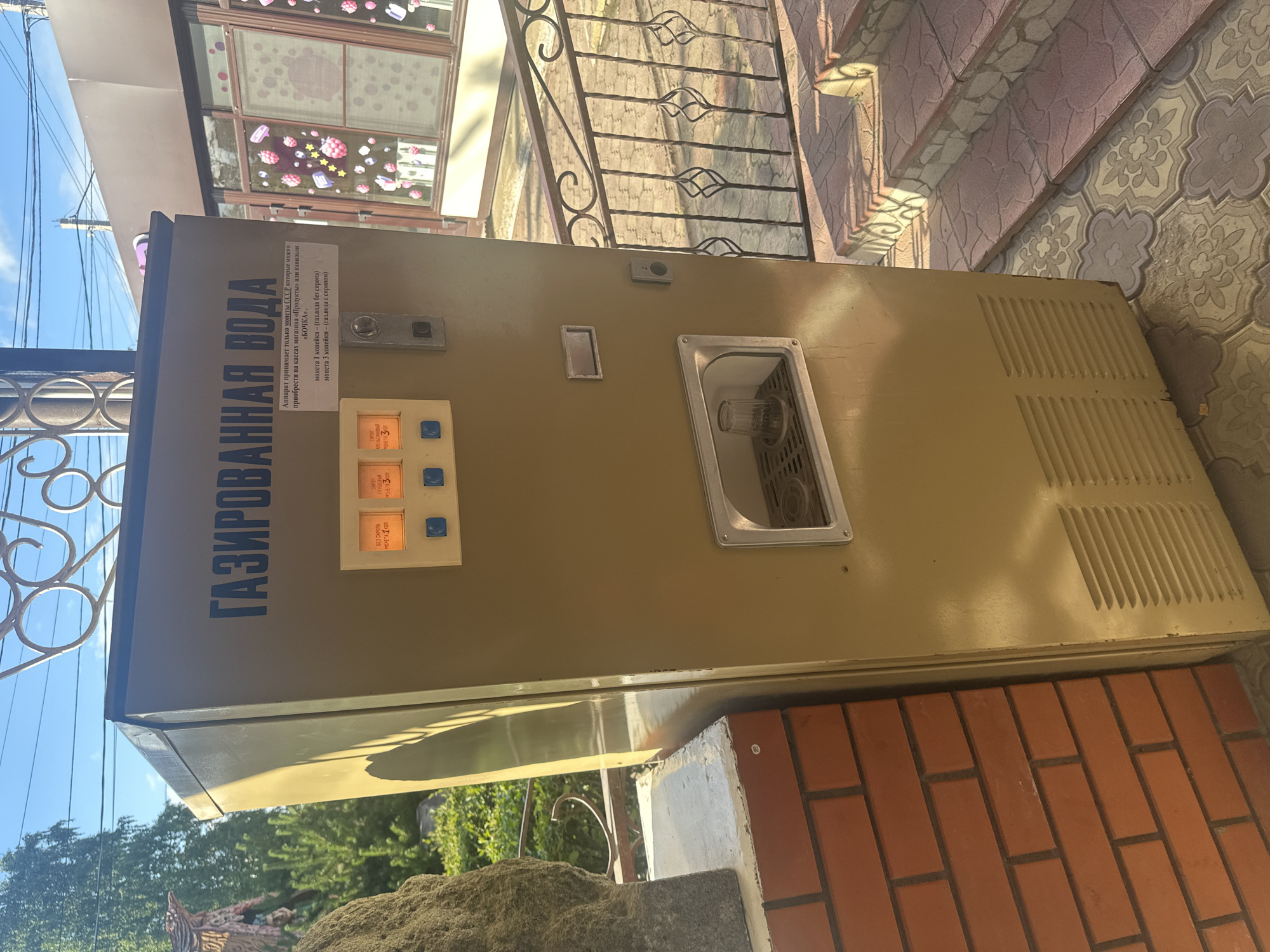 This is a full shot, eye-level, outdoor photograph of a vintage soda vending machine located in Kablukovo, Russia. The machine is tan and appears to be made of metal. It has a large sign on the front with Russian text that reads "GAZIRVANNAYA VODA," meaning "sparkling water." Below the sign are three illuminated buttons with prices displayed in kopecks: "3 kopecks - plain water," "3 kopecks - water with syrup," and "5 kopecks - water with syrup." To the right of the buttons is a slot for inserting coins and a dispenser area where a glass is visible. The machine is situated on a tiled patio with a decorative metal railing and steps leading up to it. To the left of the machine is a brick wall and a large, textured rock. In the background, trees and a building with decorated windows can be seen. The lighting suggests it is a sunny day.