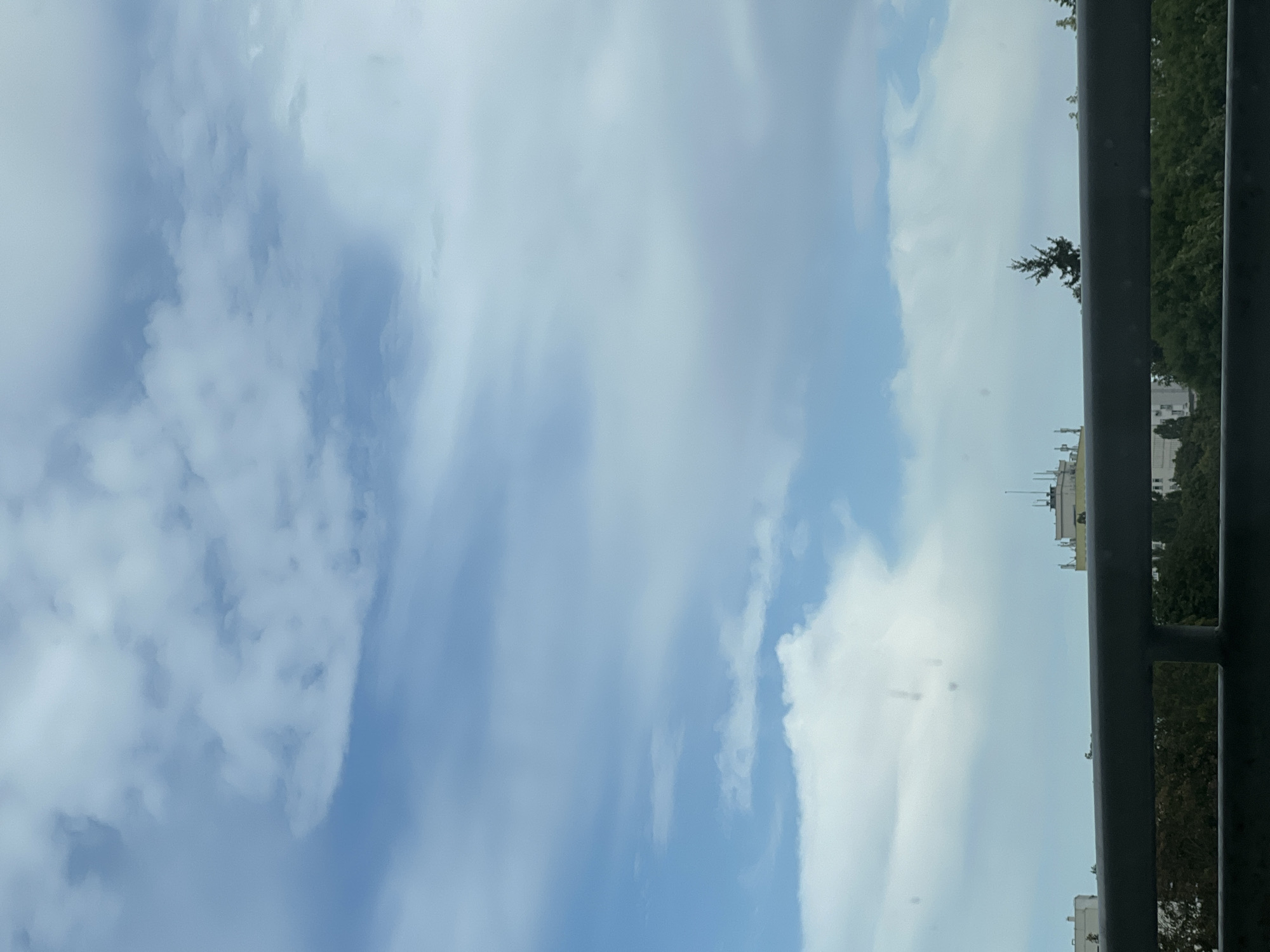 The image depicts a view of the sky and a cityscape through a railing. The sky is filled with a mix of light and dark clouds against a blue backdrop. In the middle ground, several buildings are visible, with one prominent structure featuring multiple antennas on its roof. A lone evergreen tree stands out against the skyline. The foreground is dominated by a dark gray railing. The scene appears to be set in a city, likely Warsaw, Poland, given the architectural style of the buildings and the presence of the evergreen tree, which is common in the region. The overall atmosphere suggests an overcast or partly cloudy day.