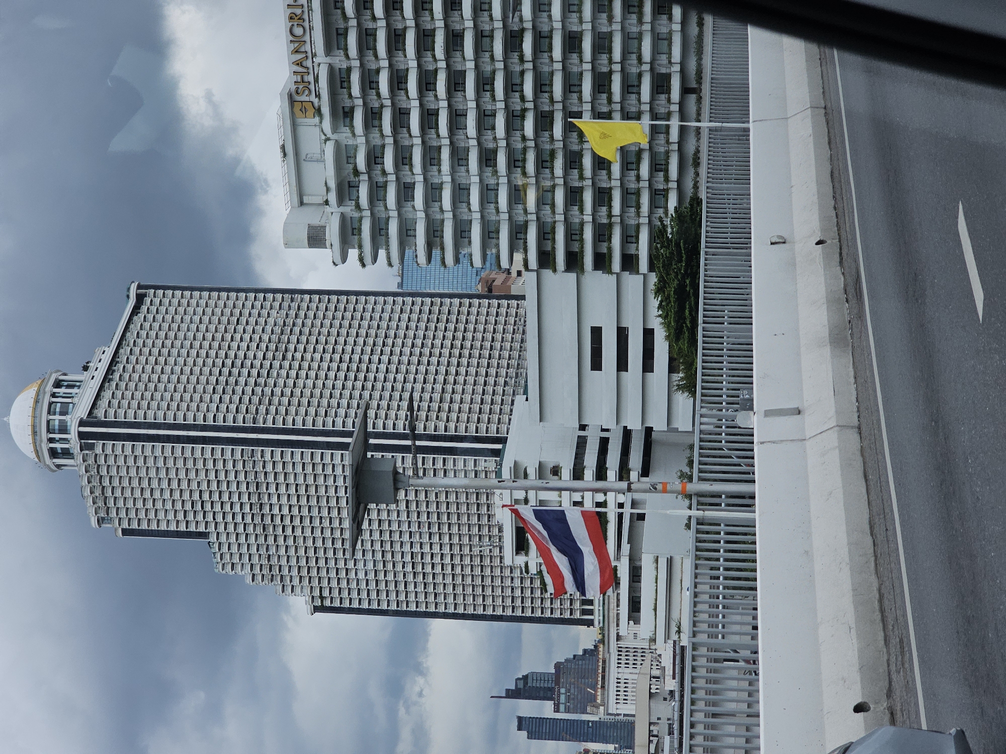 The image shows a view of the Bangkok skyline from a street level perspective, likely on a bridge or elevated road. In the foreground, a concrete barrier separates the road from a metal fence. The road is asphalt with white lane markings.
Dominating the mid-ground and background are several large, modern buildings. To the left is a distinctive tall skyscraper with a stepped design, covered in a grid of balconies and topped with a golden dome. To the right is another large white building with a facade of curved balconies and the "SHANGRI-LA" logo visible near the top. The Shangri-La hotel is located in Bangkok, Thailand. The sky is overcast and cloudy.
Two flags are visible. A Thai flag (red, white, blue, white, red stripes) is flying from a tall pole in front of the stepped skyscraper. A plain yellow flag is flying from a shorter pole in front of the Shangri-La building.
In the far background, other skyscrapers of varying heights and architectural styles are visible, suggesting a dense urban environment. The overall scene depicts a section of Bangkok's commercial and hotel district under a cloudy sky. There are no visible people or vehicles in motion in the immediate scene.