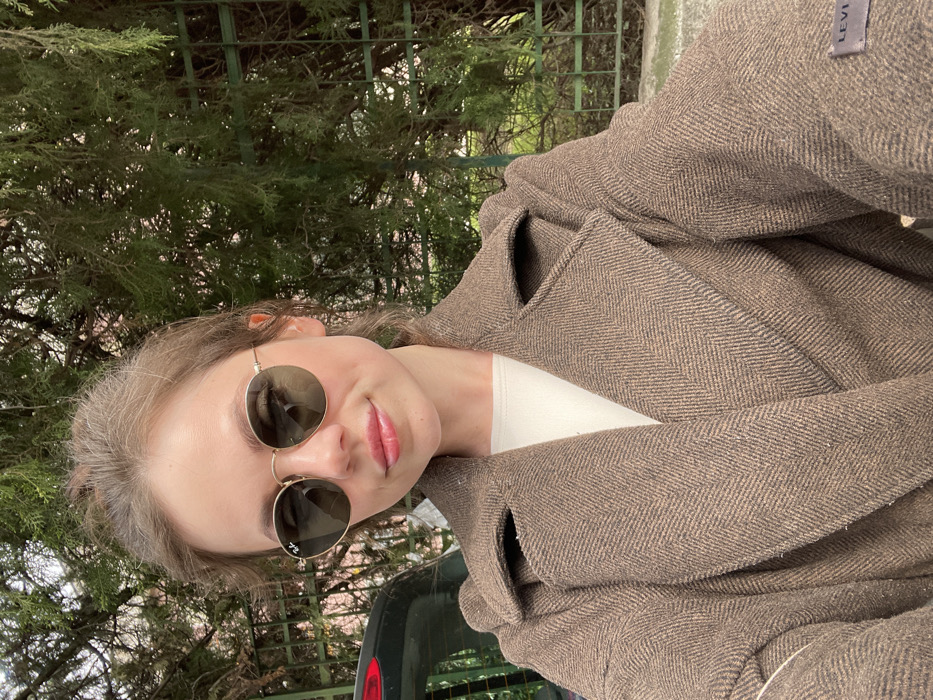 A female individual is depicted in a medium shot, facing the camera, outdoors in Cava de' Tirreni, Italy. She has brown hair pulled back and is wearing round, gold-rimmed Ray-Ban sunglasses with dark lenses, the brand logo visible on the right lens. Her lips are visible and exhibit a light pink or gloss application. She is dressed in a white inner garment beneath a thick, brown herringbone-patterned overcoat with wide lapels. A 'LEVI'S' brand label is present on the left sleeve of the overcoat. The background features dense green foliage from trees or shrubs, partially obscuring a dark green metal fence or trellis on the right. On the left, the rear section of a dark-colored vehicle, including a red taillight, is partially visible. A light-colored paved surface is visible at the bottom of the frame.