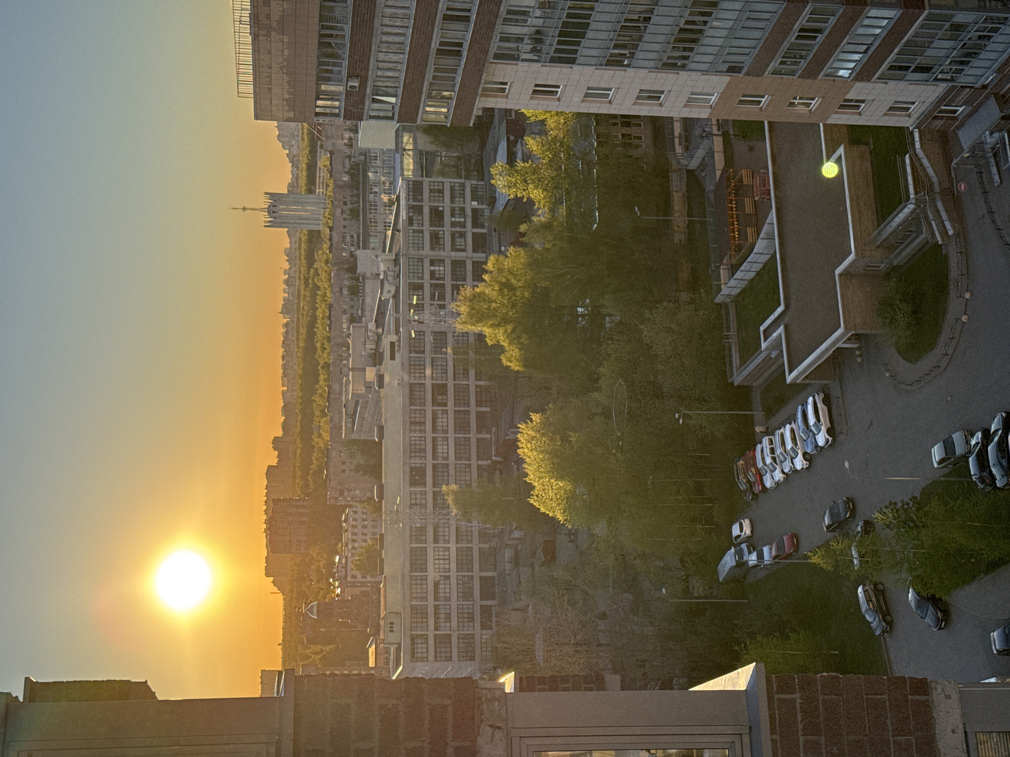 The image is a high-angle shot of an urban landscape at sunset. In the foreground, there is a portion of a building with a brick facade. The middle ground shows numerous buildings of varying heights, interspersed with trees. A line of parked cars is visible. The background shows the sun setting, casting a golden light over the scene. A tall communications tower stands in the distance. The location is Unknown, Unknown.