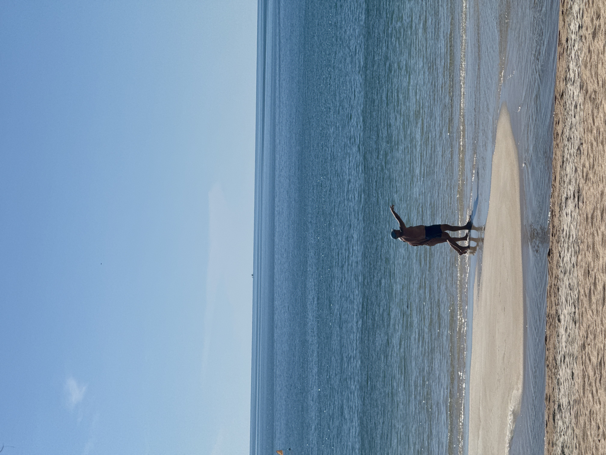 A person stands at the edge of the sea, pointing towards the horizon. The person is wearing dark shorts and a hat. They are walking into the shallow water on a sandy beach. The sea is a deep blue, with a clear sky above. The beach is made of light brown sand. There are small waves breaking on the shore. In the distance, a faint silhouette of a ship can be seen on the horizon.