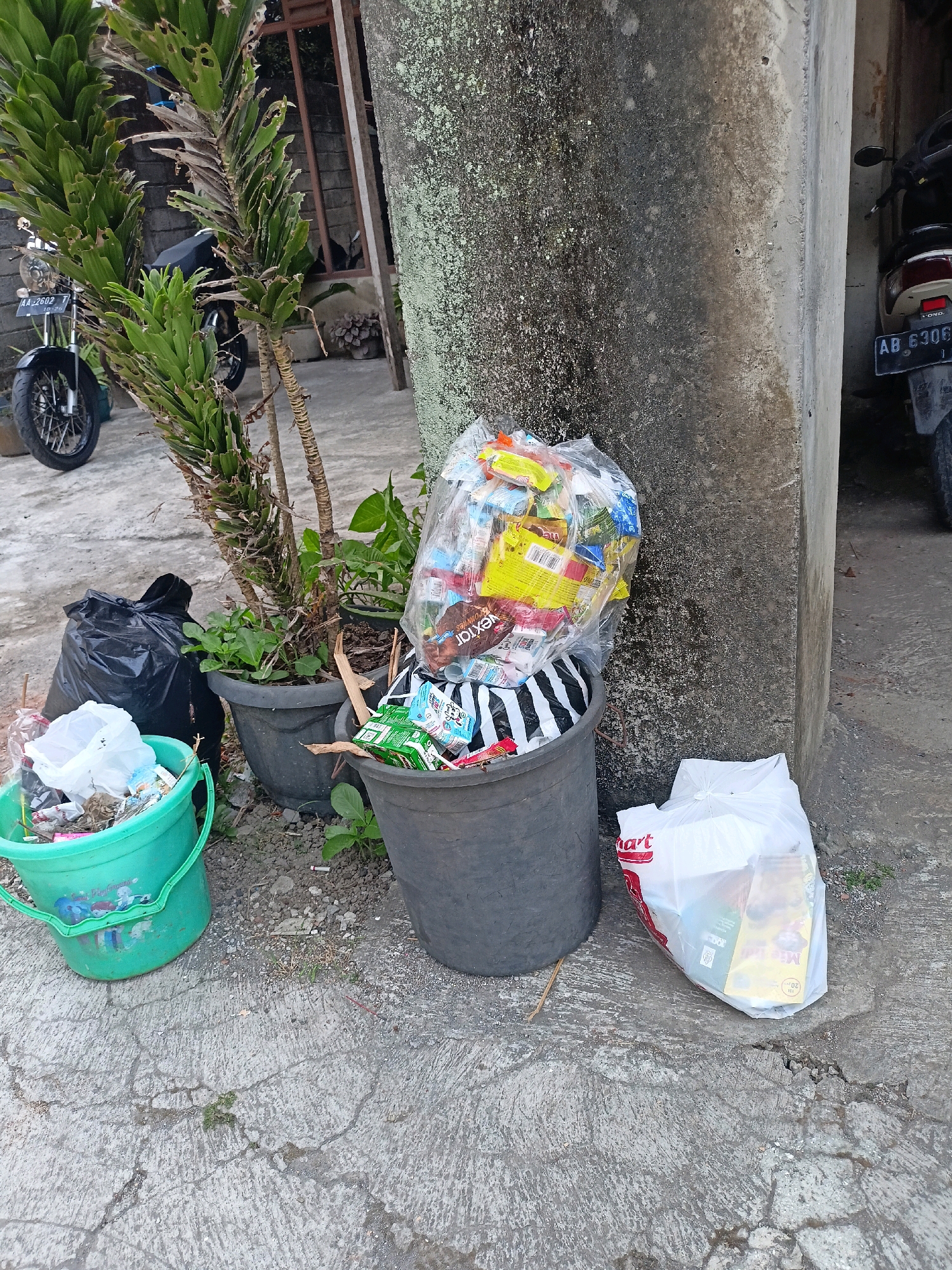 In the city of Payaman, Indonesia, a scene of scattered refuse is depicted. A large, dark gray trash can overflows with plastic packaging, food wrappers, and other assorted waste, some of which has spilled onto the cracked concrete ground. A smaller, bright green plastic bucket sits to the left, also containing a mixture of trash and plastic bags. Beside it, a large black trash bag stands upright, its contents concealed. To the right, another white plastic bag, bearing the red "Hart" logo, is filled with discarded items, including what appears to be food packaging. A tall, textured concrete pillar, covered in patches of green lichen, anchors the right side of the frame. Behind the trash cans and bucket, a motorcycle is partially visible, along with a lush green plant in a dark pot. Further in the background, a structure with a window and a wooden frame is apparent. The overall environment suggests an outdoor area, possibly a roadside or a residential courtyard, with a focus on the accumulation of waste.