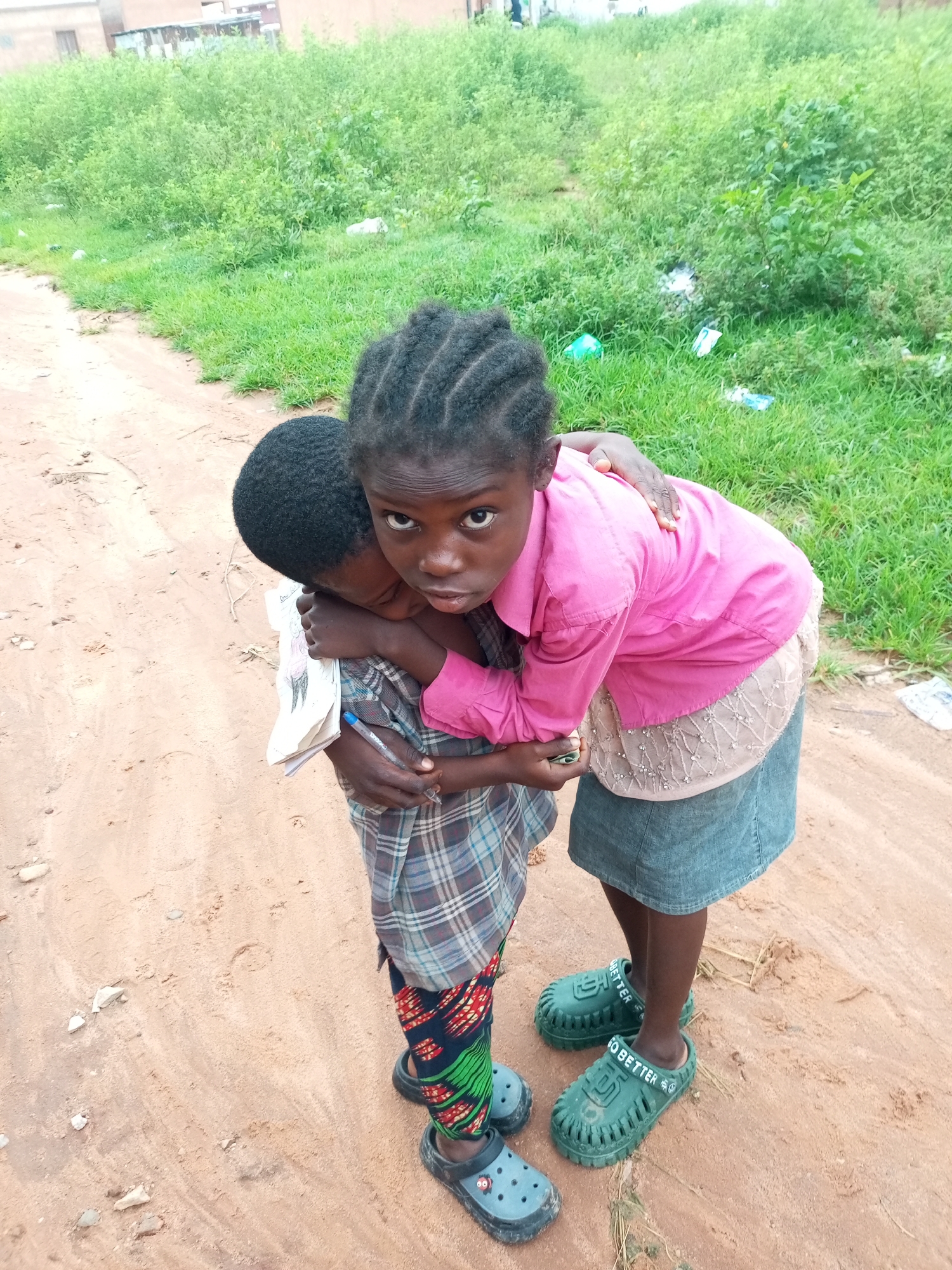In Tempure, Nigeria, two young Black children embrace on a dirt path bordered by lush green vegetation and distant buildings. The child on the right, facing the camera with a direct gaze, wears a bright pink collared shirt and a denim skirt. Their hair is styled in braids. The child on the left, whose back is partially to the camera, is embraced by the other. This child wears a plaid shirt and a patterned fabric around their lower legs, with dark blue clogs on their feet. The child on the right wears green clogs with "GO BETTER" embossed on them. The background shows a dense area of green plants and some scattered litter. The path is sandy with some small stones.