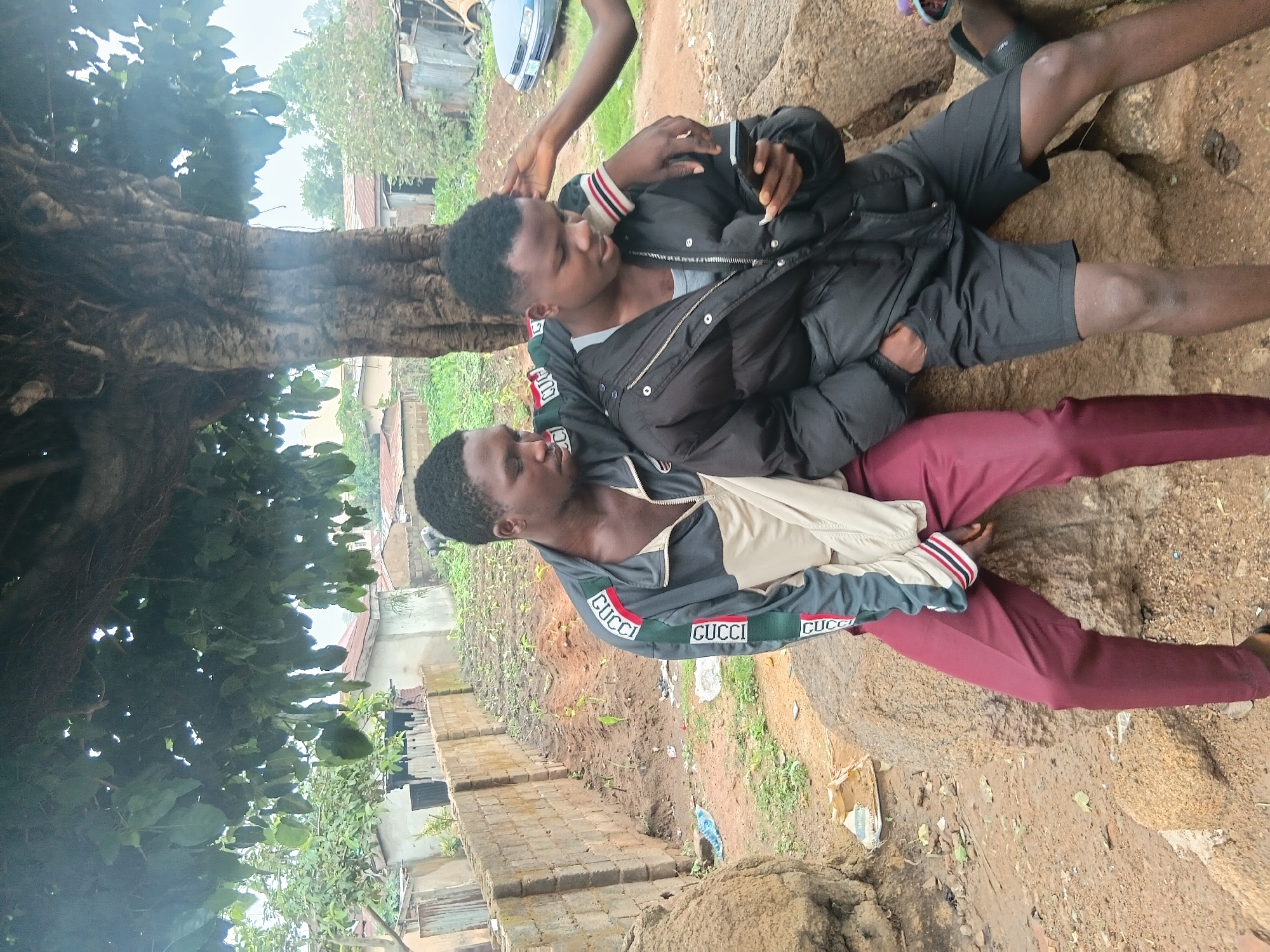 In Jos, Nigeria, two young men are seated on large rocks under a mature tree. The man on the left is wearing a gray and cream-colored jacket with green and red trim, and maroon pants. The man on the right is wearing a black puffer jacket over a gray shirt, and black shorts. He is holding and looking at a smartphone. The arm of a third person is resting on his shoulder. The background shows a dirt path, a brick wall, and several buildings with corrugated metal roofs, typical of a residential area in Jos. Lush green vegetation is visible throughout the scene.