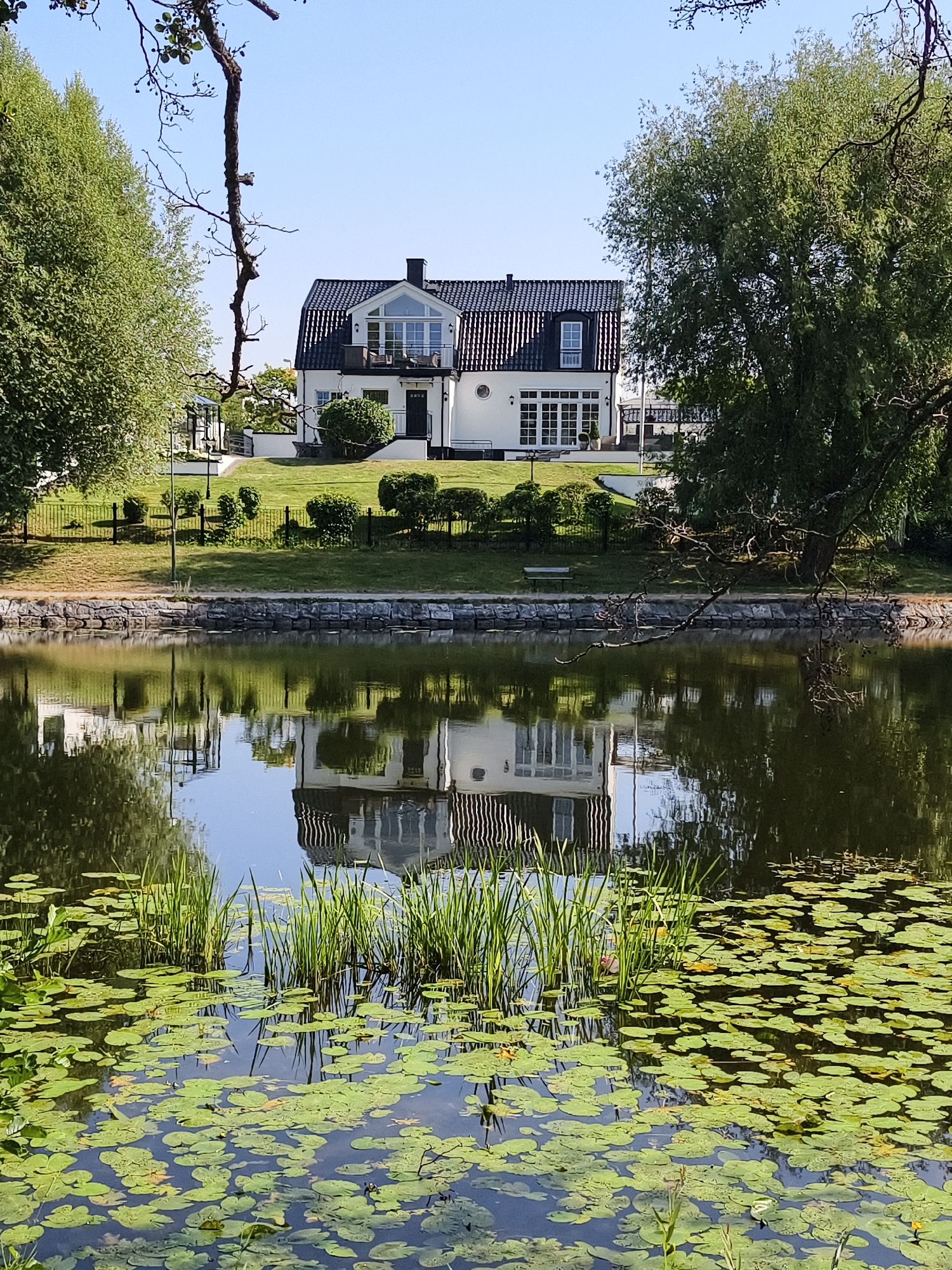 The image depicts a modern white house with a black roof and multiple windows, set against a clear blue sky. The house is bordered by a well-maintained lawn and several trees. In the foreground, a body of water with lily pads and reeds reflects the house and the surrounding trees. The scene appears to be a serene residential area, possibly a lakeside or riverside property in Linköping, Sweden, given the architectural style and vegetation. No people are visible in the image.