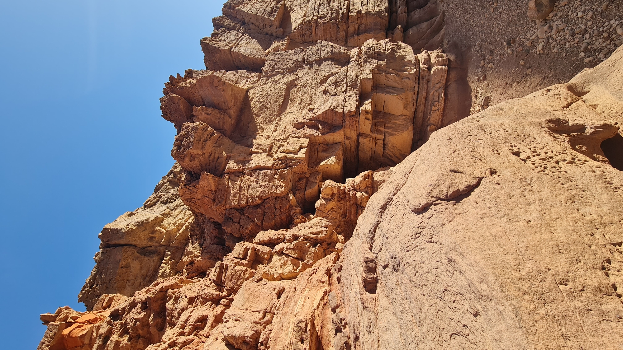 The image depicts a close-up view of a rocky terrain, likely a geological formation. The rocks are predominantly brown and orange, with varied textures, ranging from smooth surfaces to fractured and layered formations. The top of the image shows layered rocks. Some rocks have small holes. The sky is visible in the upper-left corner, appearing as a clear, bright blue. The image was taken in Hevel Eilot Regional Council, Israel.