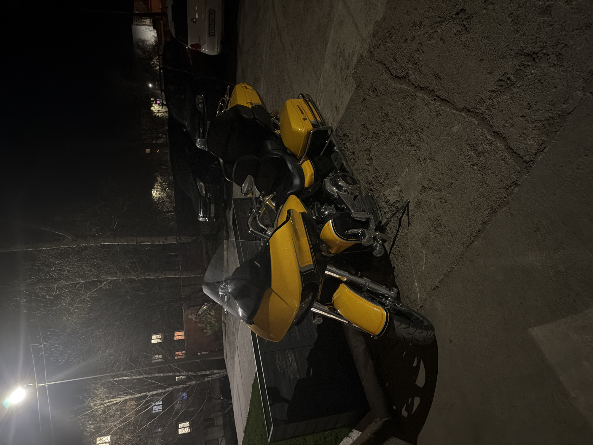 A yellow and black motorcycle is parked on a paved surface outdoors at night. The motorcycle has saddlebags and a windshield. In the background, there is a building with illuminated windows and trees. A white car is partially visible in the background. The location is the city of Votkinsk, Russia.