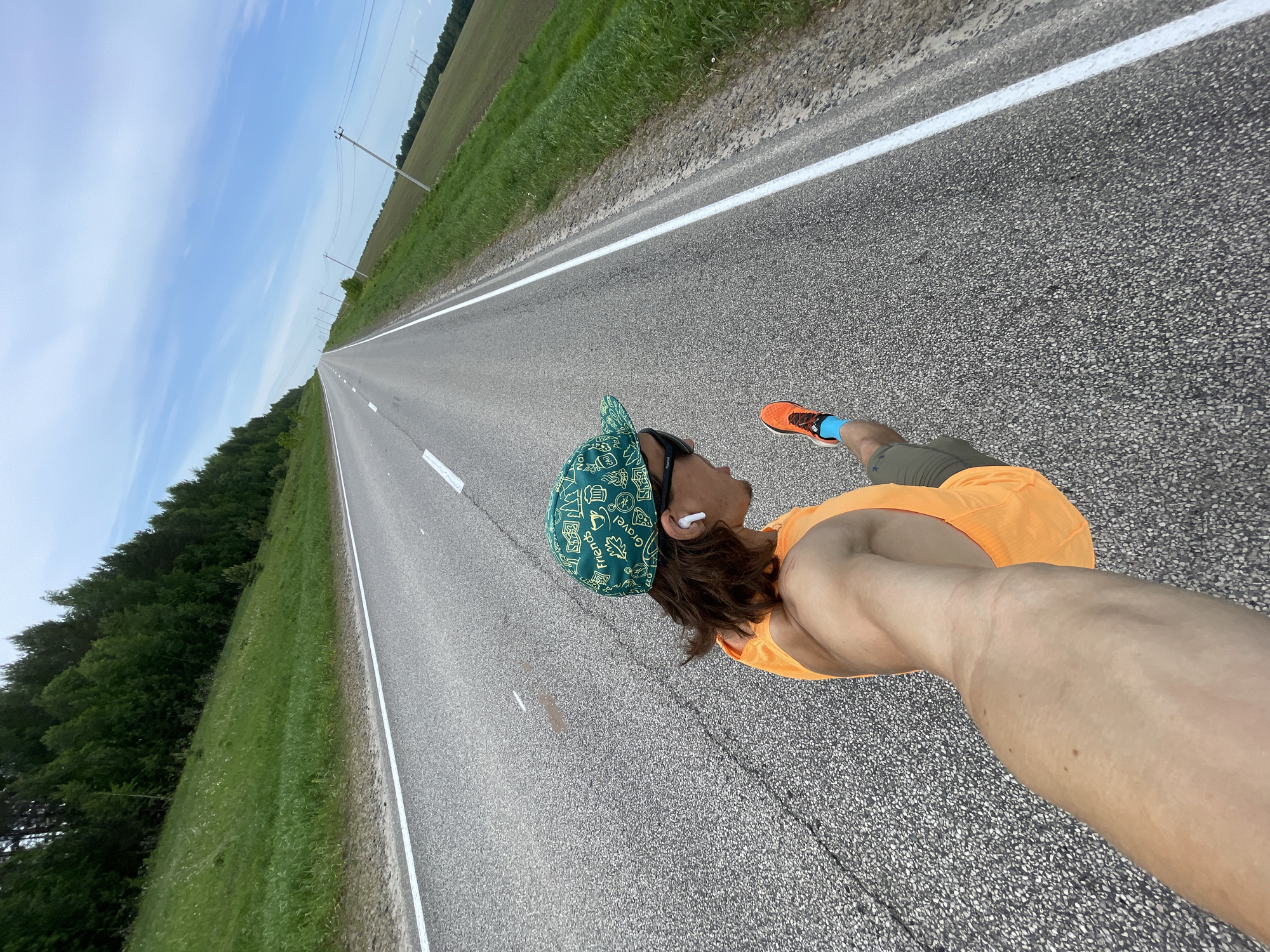 A person is running on a straight asphalt road in Перевозное, Russia. The road has a solid white line down the center. To the left of the road is a green grassy area bordering a forest of trees. To the right is a similar green area and field. The person is wearing an orange shirt, green hat, running shoes, and sunglasses. They have a wireless earbud in their ear. The sky is blue with scattered clouds. Utility poles are visible along the roadside in the distance.