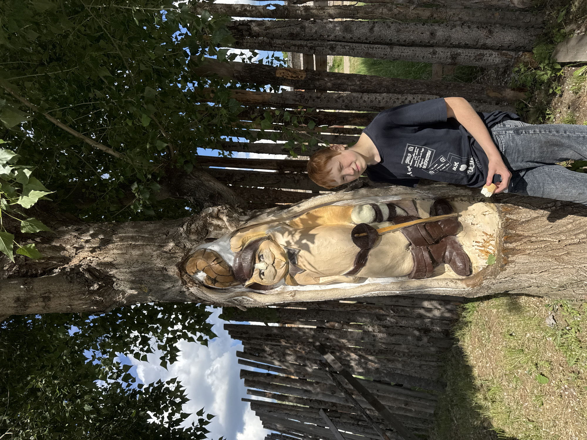 A boy is standing next to a tree carving in the city of Chaikovsky, Russia. The boy is wearing a dark t-shirt and blue jeans. He is holding something white in his hand. The carving is of a man in a hat, laying down with a sword on top of him. The tree is next to a wooden fence, and there are trees overhead. The sky is visible between the trees.