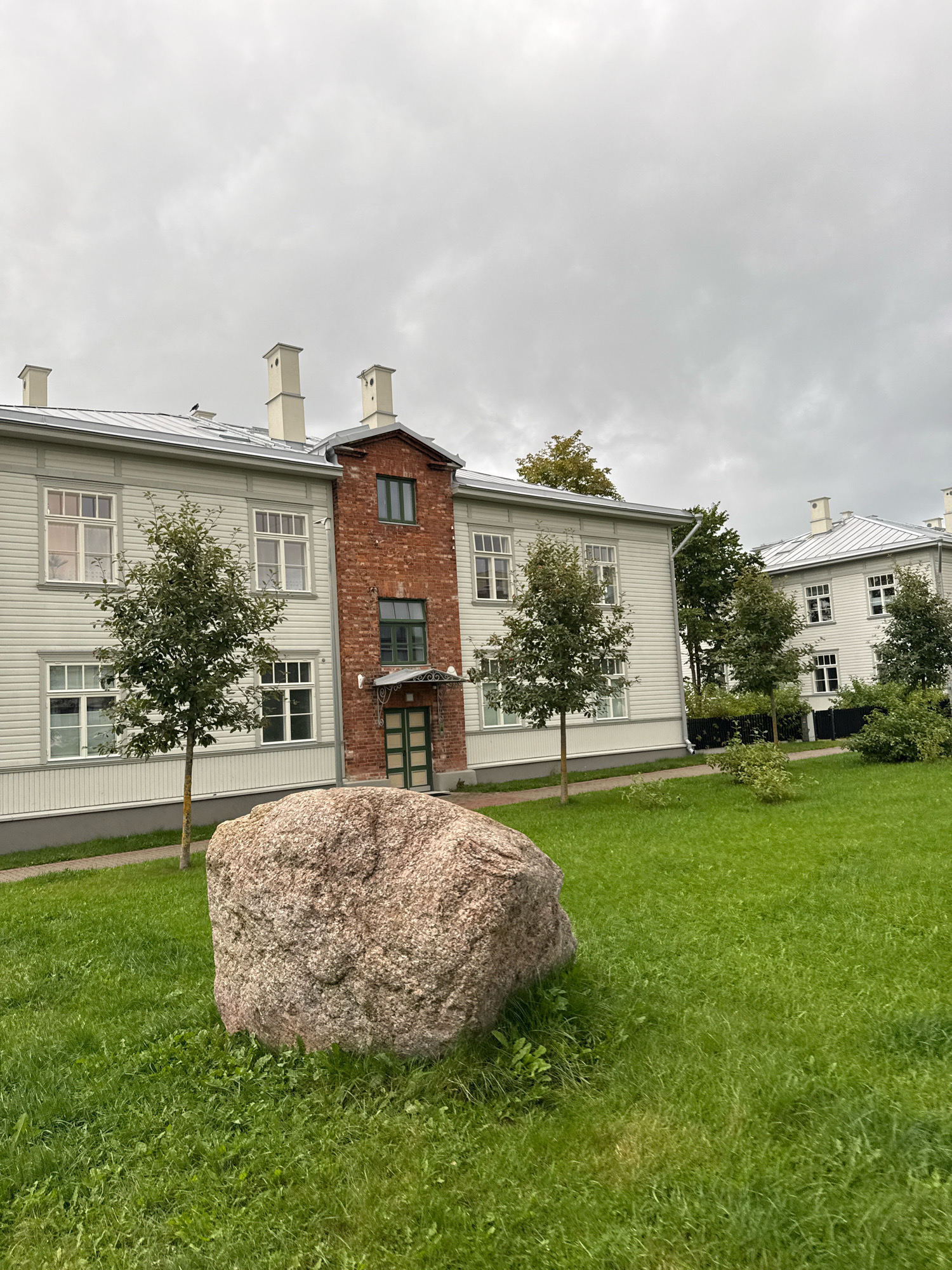 The image depicts an exterior view of a building complex located in Tallinn, Estonia. The architecture suggests a residential or institutional setting, possibly a former barracks or a school. The buildings are low-rise, featuring light-colored horizontal siding, white-framed windows, and prominent brick-clad sections with decorative brickwork around entrances. Multiple chimneys with rectangular caps dot the roofs.
In the foreground, a large, rough-textured boulder rests on a lush green lawn. Several young trees with green foliage are strategically planted in the lawn area, suggesting a landscaped environment. The sky is overcast and cloudy, contributing to a subdued lighting condition. The scene is devoid of any human presence or activity. The overall impression is one of a well-maintained, quiet environment, likely during daytime under a cloudy sky.