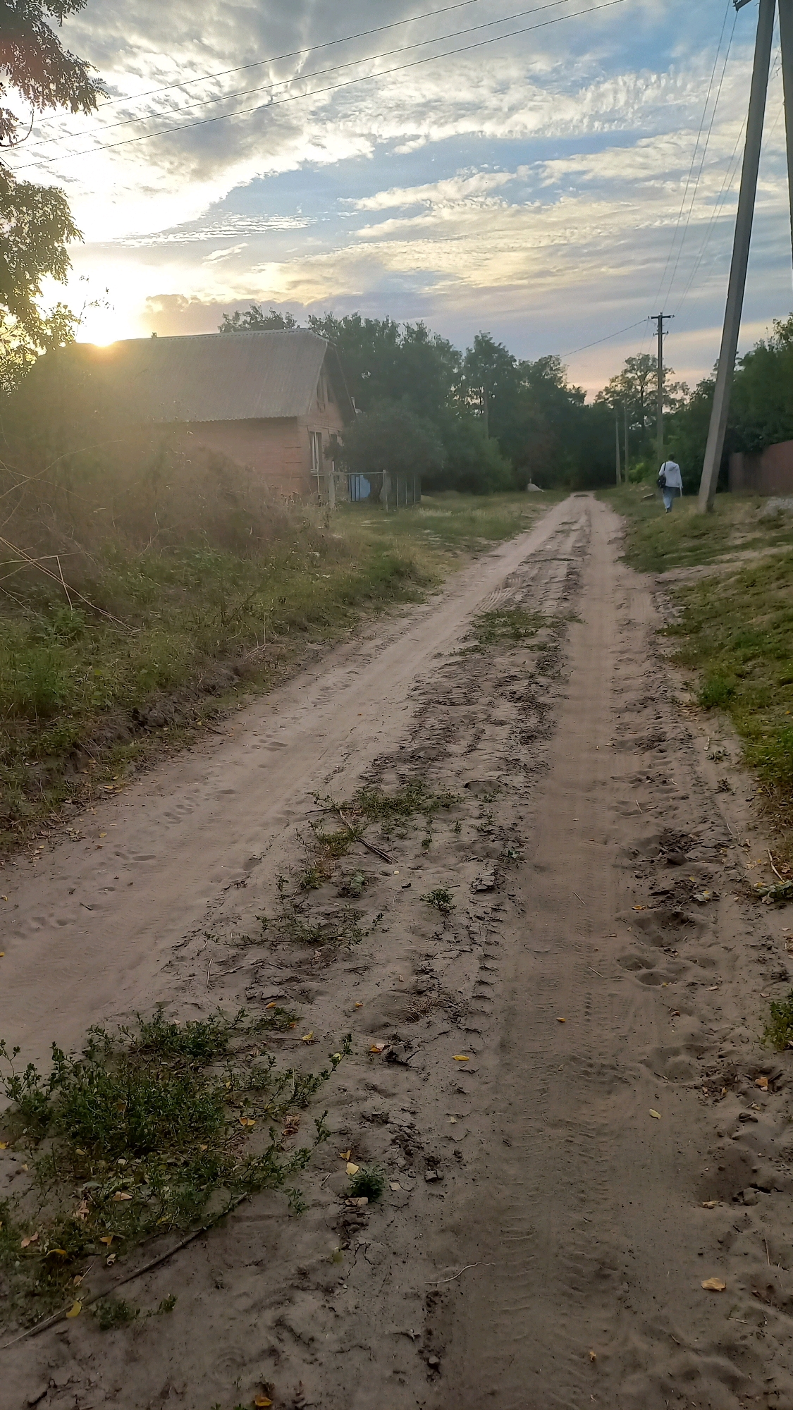 A dirt road stretches into the distance, flanked by overgrown grass and trees. A small, brick house with a corrugated metal roof sits to the left, partially obscured by a sun flare. In the distance, a person walks away from the camera, towards a utility pole and a low fence. The sky above is a mix of blue and white clouds, with the sun peeking through on the left. The scene appears to be in a rural or suburban area of Klyntsi, Ukraine, likely during the late afternoon or early evening, given the lighting.