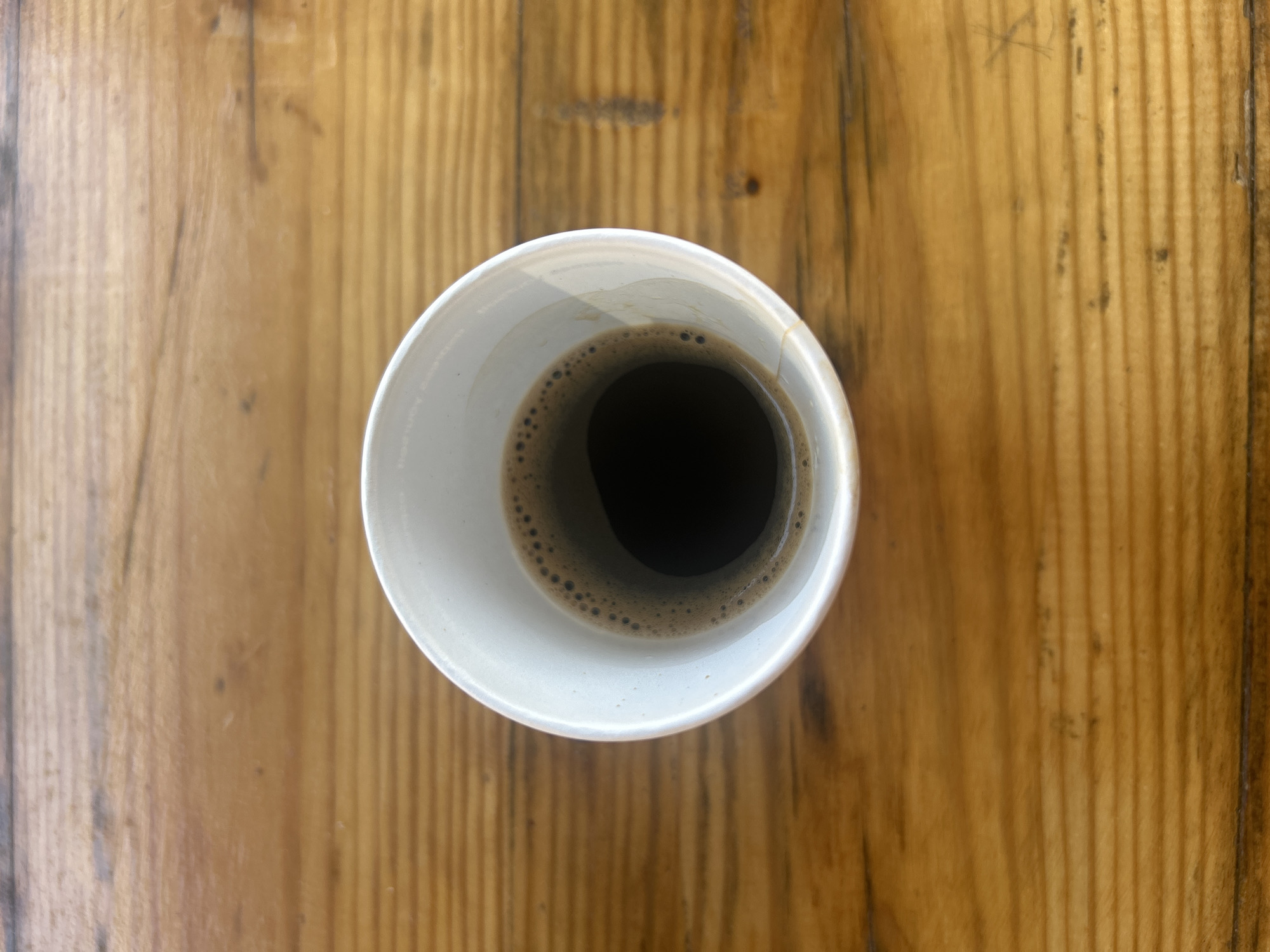 A close-up, top-down view shows a white paper cup of dark coffee, viewed from above. The coffee has a foamy layer around the rim. The cup rests on a polished wooden surface with visible grain. The image does not provide information about the city of Martinovo, Bulgaria, or any people, actions, or interactions.