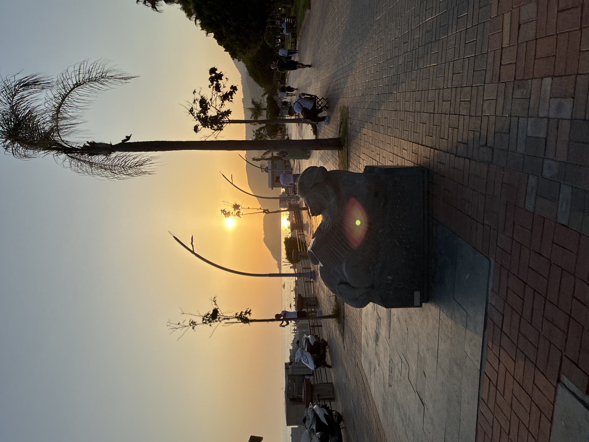 The image depicts a sunset view along the Alanya promenade in Turkey. The foreground features a textured stone sculpture, possibly depicting a reclining figure, placed on a paved walkway composed of interlocking bricks and larger stone slabs. To the left, several parked scooters, including a white one, are visible. Beyond the promenade, the Mediterranean Sea stretches out, with a distant hillside and a small town visible on the horizon. Several individuals are walking and sitting along the promenade. A man stands near a tree, seemingly taking a photo, while others stroll further down the path. A tall palm tree stands prominently on the right side of the frame, casting a shadow on the walkway. The sky is a gradient of soft blues and oranges, indicating the sun is low on the horizon, creating a warm, diffused light that reflects off the water. The overall atmosphere is serene and suggests an early evening scene on a popular public walkway.