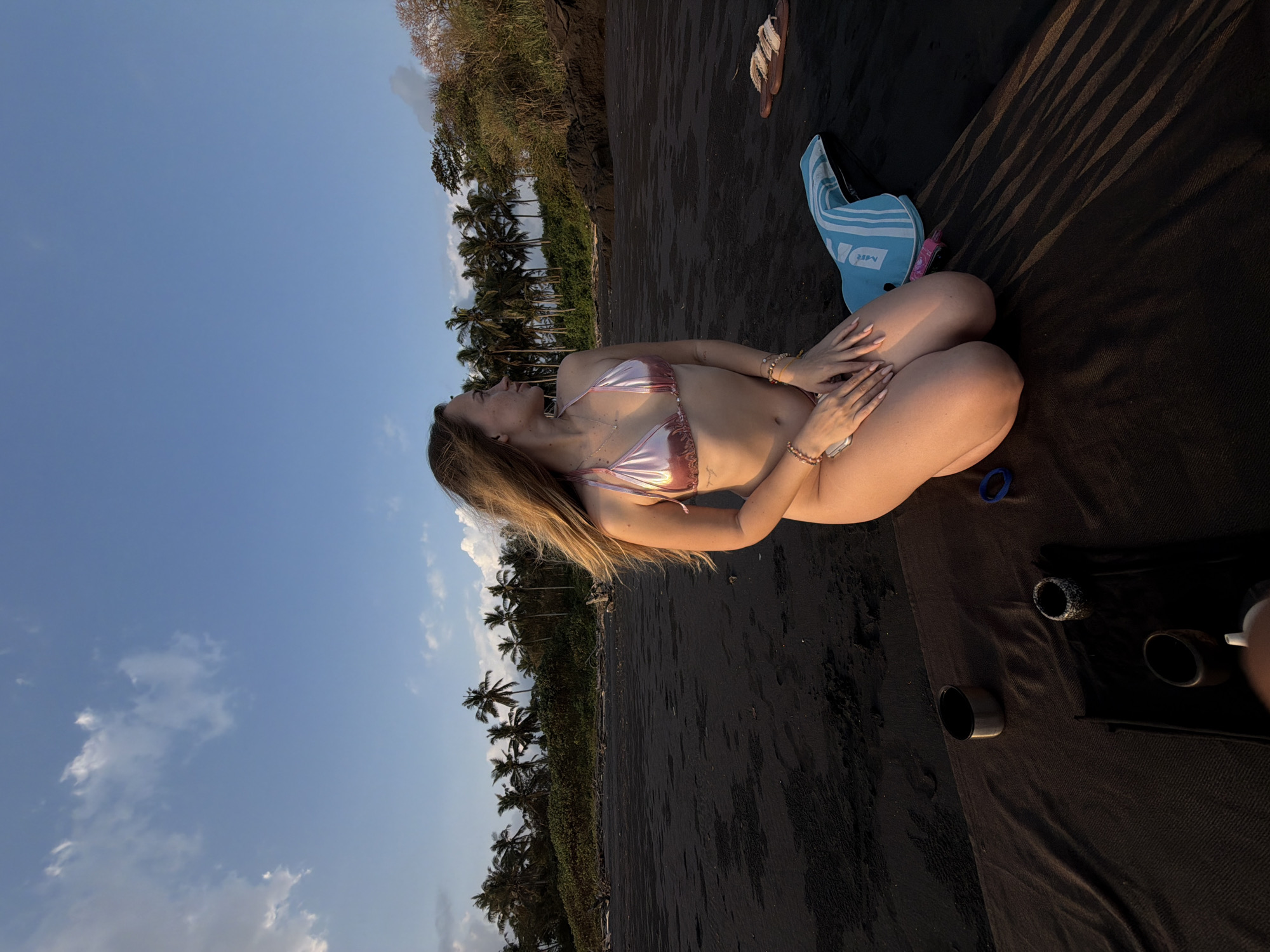 A young woman with blonde hair is kneeling on a black sand beach in Selemadeg Timur, Indonesia. She is wearing a pink bikini and looking to her right, away from the camera. The beach is lined with palm trees and lush vegetation. In the foreground, there is a dark blanket with a few small cups or bowls placed on it, as well as a blue and white striped towel and a pair of flip-flops. The sky is blue with scattered clouds. The scene appears to be captured during the late afternoon or early evening, as indicated by the warm lighting and long shadows.