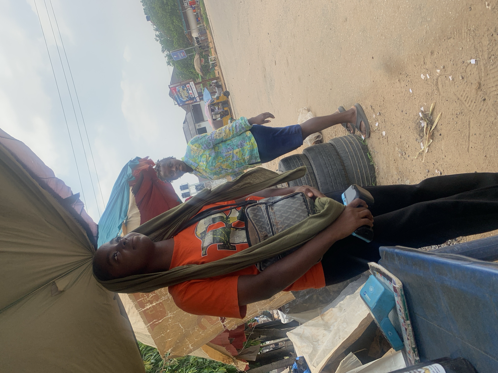 The image depicts an outdoor scene on what appears to be a street in Abuja, Nigeria. In the foreground, a young Black person wearing an orange t-shirt, black pants, and a green headscarf is standing. They are holding a blue mobile payment device in their right hand and have a patterned crossbody bag. To their left, a blue table holds another mobile payment device and some papers. Behind the foreground figure, a second Black person, dressed in a light blue and green patterned shirt and dark blue skirt, is standing and looking towards the camera. They are wearing flip-flops.
The background shows a street with various elements suggesting a commercial or roadside area. A few vehicles, including a yellow auto-rickshaw, are visible further down the street. There are signs, including one that reads "SUPERNATURAL EDGE." Stacks of tires are present near the foreground figures, and overhead, a makeshift canopy provides shade, constructed from what appears to be tarps and fabric. The ground is a mix of sand and dirt, with some scattered debris. The sky is overcast. The overall impression is of a street-side scene, possibly a marketplace or a bus stop, in an urban environment in Abuja.