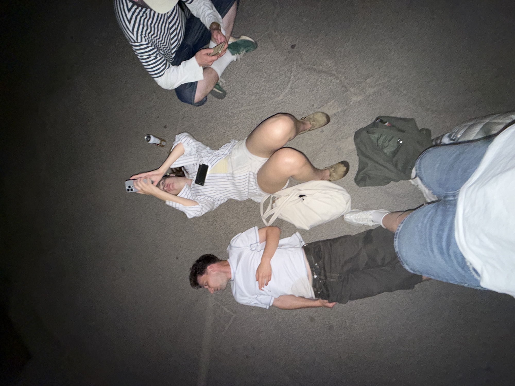 Four people are on a paved surface, possibly a street, in Cukarica Urban Municipality, Serbia. A person sits in the upper center, wearing a striped shirt and shorts, looking at a phone. A person lies on their back, wearing a striped shirt and shorts, holding a phone. A person lies on their side, wearing a white t-shirt and dark pants. The fourth person's legs are visible in the lower right, wearing blue shorts and a white shirt. A can is near the person lying on their back. A white bag and green jacket are also on the ground.