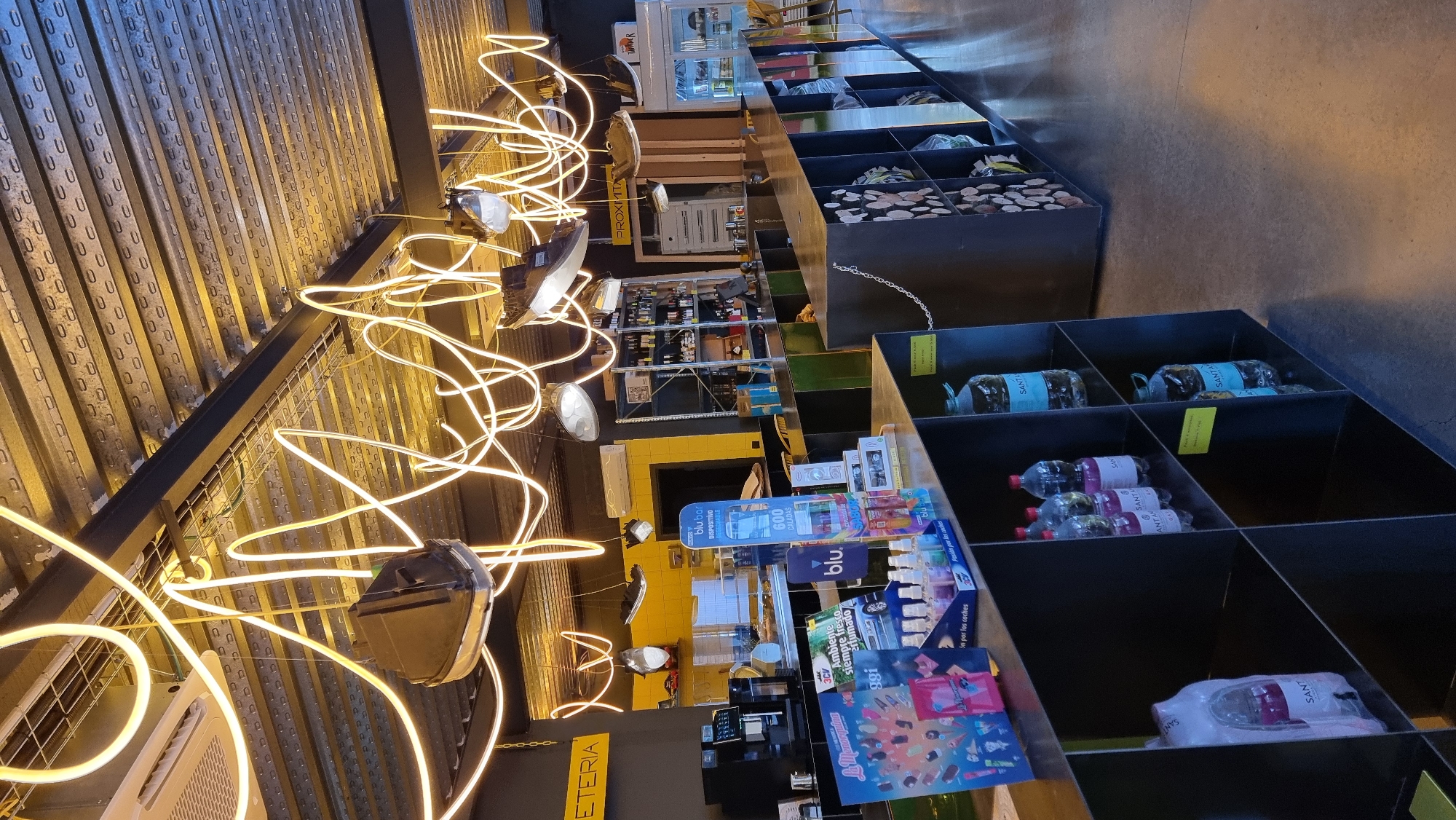 The image shows the interior of a modern convenience store or small supermarket, possibly a "Proximiti" brand based on signage. The store features a long, dark counter with shelves below, displaying bottles of water and other items. Overhead, a unique lighting system of intertwined yellow neon tubes illuminates the space, creating a vibrant and contemporary ambiance. In the background, shelves stocked with bottles and other products are visible, along with a coffee machine and various displays of merchandise. The floor is a polished concrete. While the image depicts a commercial space in Olot, Spain, no specific people or interactions are captured.