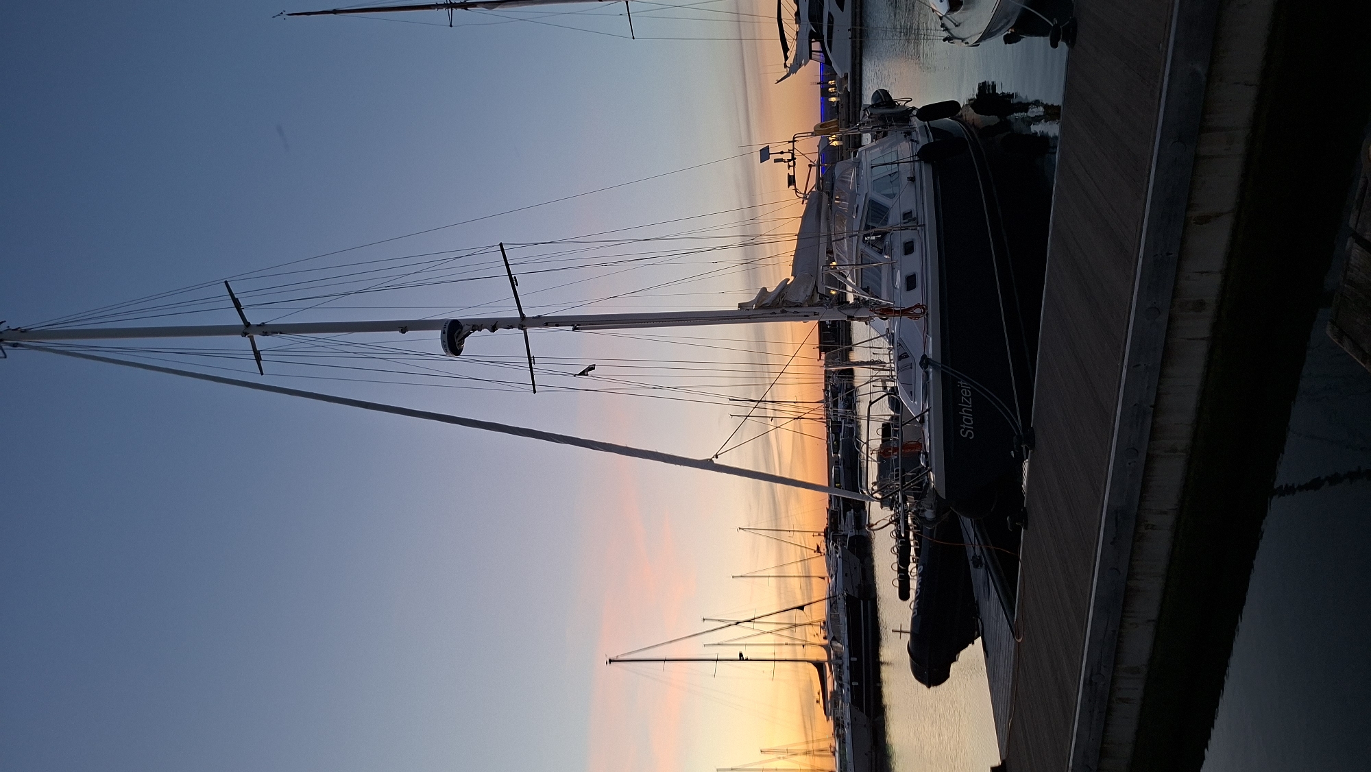 The image shows a marina in Tallinn, Estonia, at sunset. The sky is a gradient of blue, pink, and orange. Several sailboats are docked, with their masts and rigging silhouetted against the colorful sky. In the foreground, a sailboat named "Stahlzeit" is visible, tied to a wooden dock. The water reflects the lights of the marina and the sky. There are no people visible in the image. The scene is peaceful and serene.