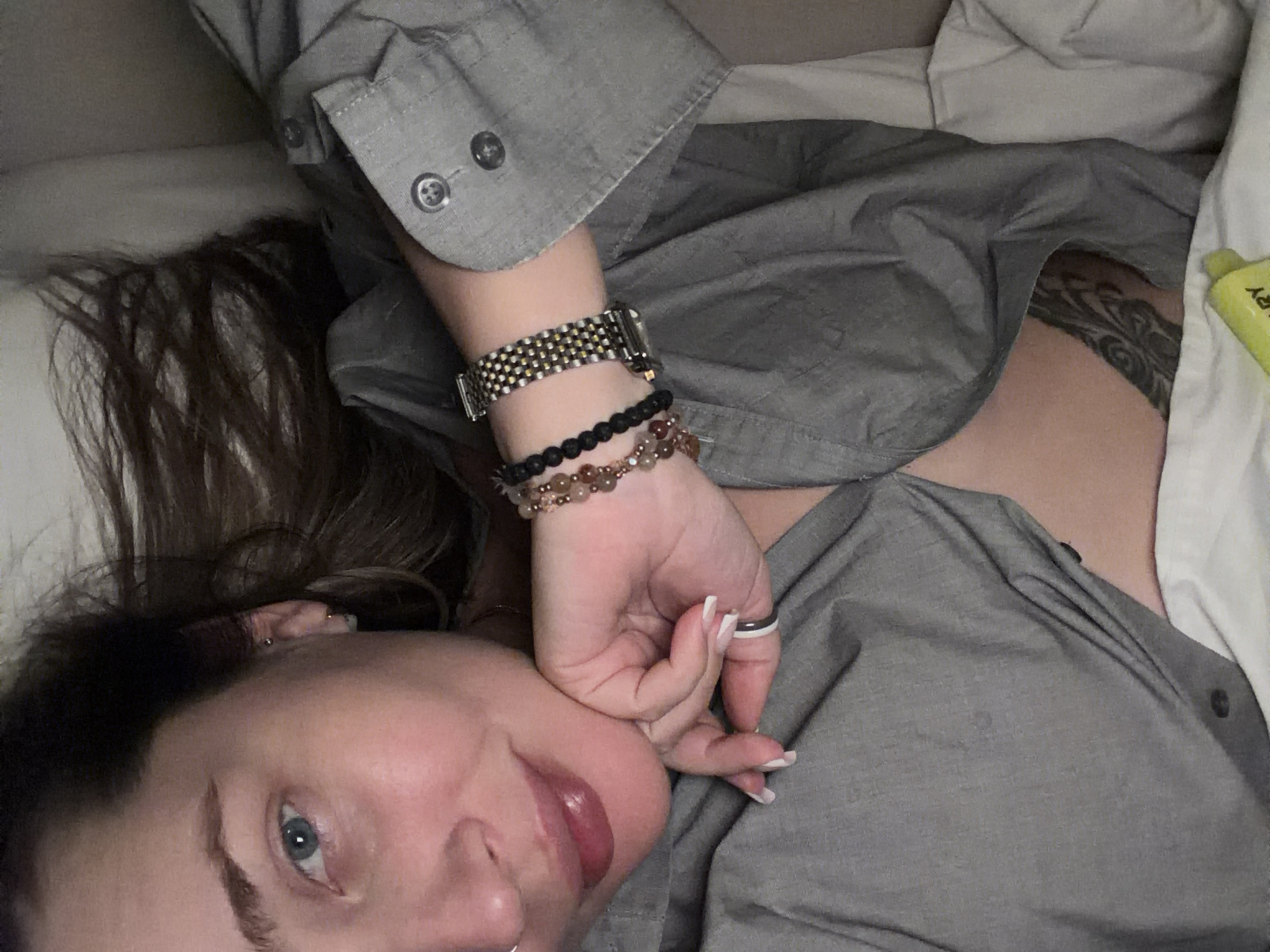 A woman lies in bed wearing a gray button-down shirt and several bracelets on her left wrist. She rests her face on her hand, looking towards the camera. Her hair is dark with lighter streaks, and her eyes are blue. A tattoo is visible on her upper chest/shoulder area. White pillows are visible in the background. There are no elements in the image that indicate the location of Moscow, Russia.