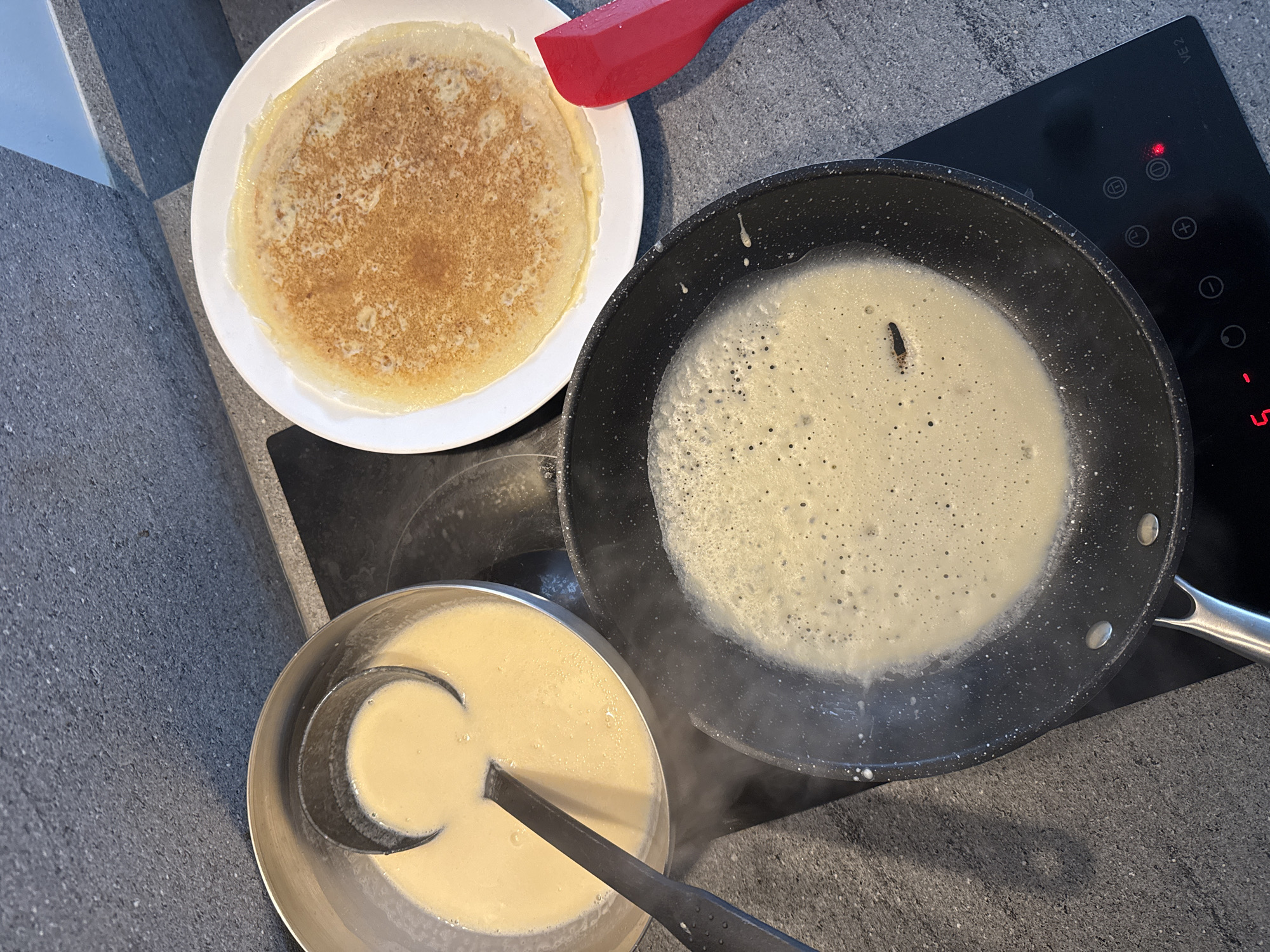 This image depicts a cooking scene, likely in a kitchen. A cooked pancake sits on a white plate, positioned next to a red spatula. Next to the plate is a black frying pan on a black stovetop containing pancake batter as it begins to cook. Steam rises from the pan. A stainless steel bowl filled with batter is to the left. Inside the bowl is a black ladle. The stovetop displays electronic controls and temperature settings. The location, Choeng Thale, Thailand, is unlikely discernible from the image alone without additional information.