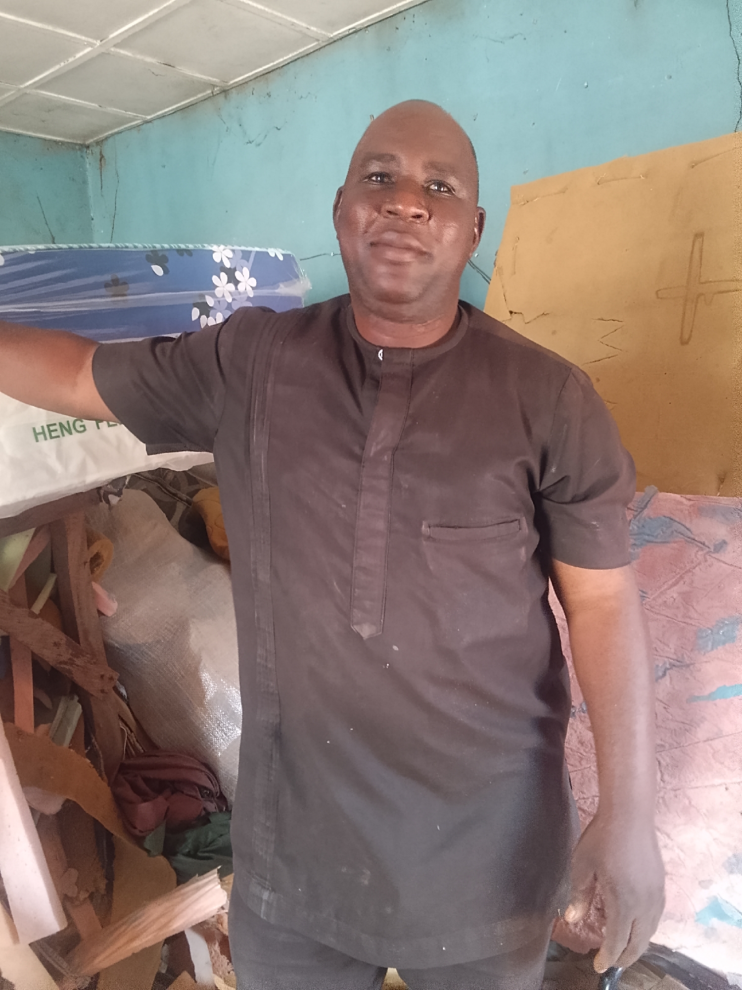 A man stands in what appears to be a workshop in Pantame, Nigeria. He is bald and wearing a short-sleeved brown shirt with black pants. He is surrounded by debris, including wood, foam, and fabric scraps. Behind him, there is a wrapped mattress with a floral pattern, a piece of cardboard with a '+' symbol, and a wall painted a light blue color. The ceiling is made of white panels. The setting suggests a small business operation involving mattresses or upholstery.
