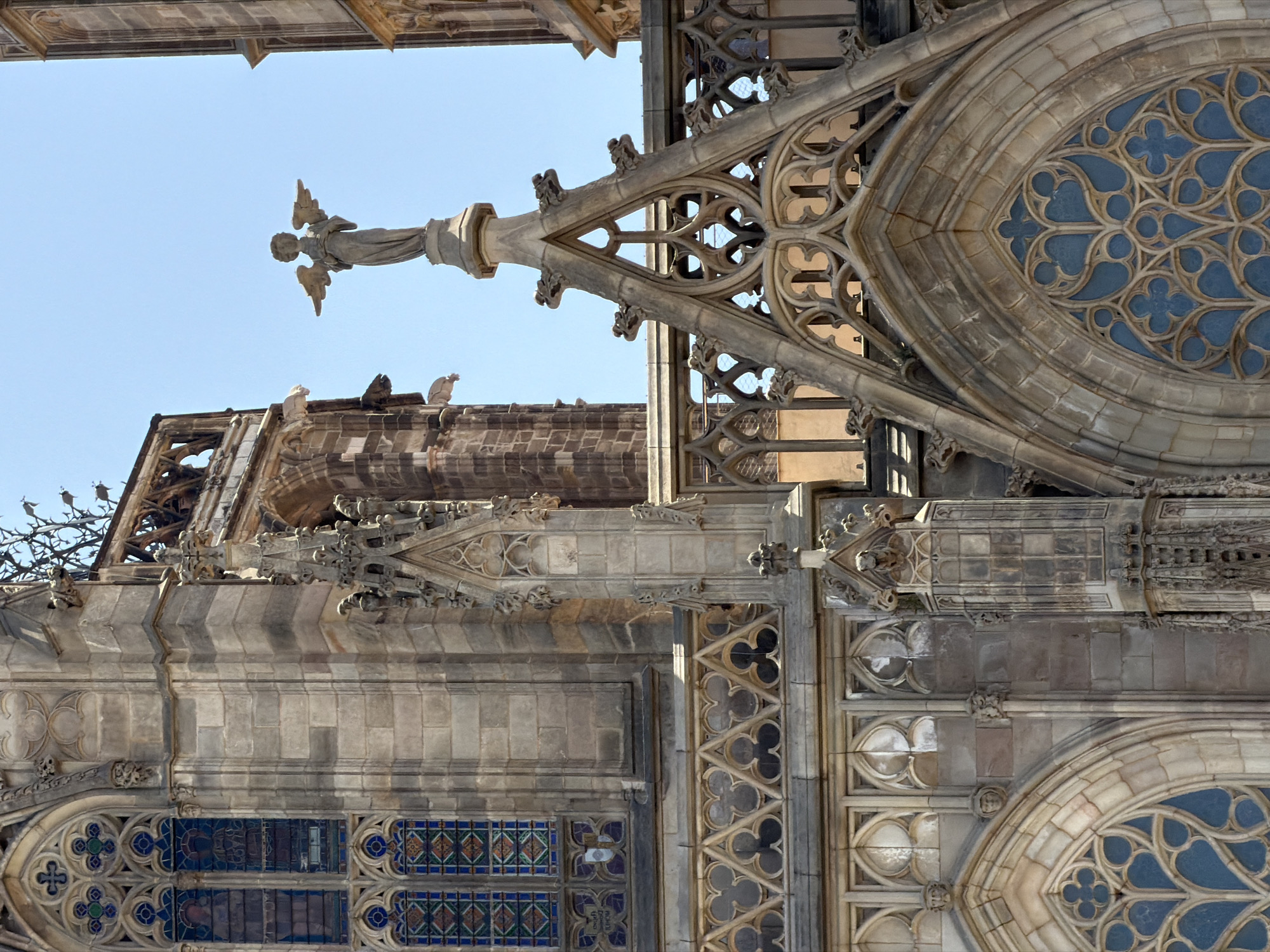 The image shows a close-up of the exterior of the Barcelona Cathedral, a Gothic Roman Catholic cathedral in Barcelona, Spain. The photograph captures architectural details of the cathedral's upper sections, including stained-glass windows, ornate stone carvings, and a statue of an angel. The statue, depicted with wings outstretched, stands atop a decorative spire. The stonework exhibits the characteristic features of Gothic architecture, with pointed arches, intricate tracery, and ribbed vaulting visible in the windows and decorative elements. The clear blue sky provides a backdrop to the detailed masonry.