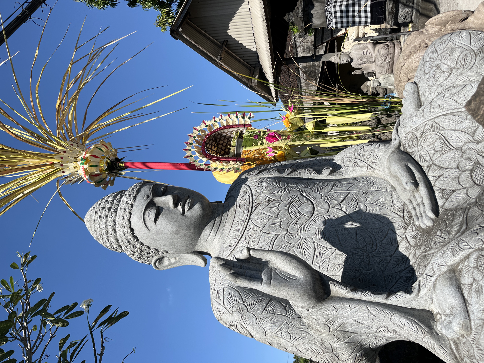 A large, grey stone statue of Buddha sits in the foreground, carved with intricate floral patterns. The statue is in a seated position with its right hand raised in a gesture of blessing. Behind the main statue, there are smaller Buddha statues, some with offerings of flowers and palm fronds. A traditional Balinese structure with a corrugated metal roof is visible in the background, along with a black and white checkered cloth draped over what appears to be a shrine. The scene is outdoors under a clear blue sky. The location is likely Sukawati, Indonesia, given the presence of Balinese cultural elements such as the offerings and the architectural style.