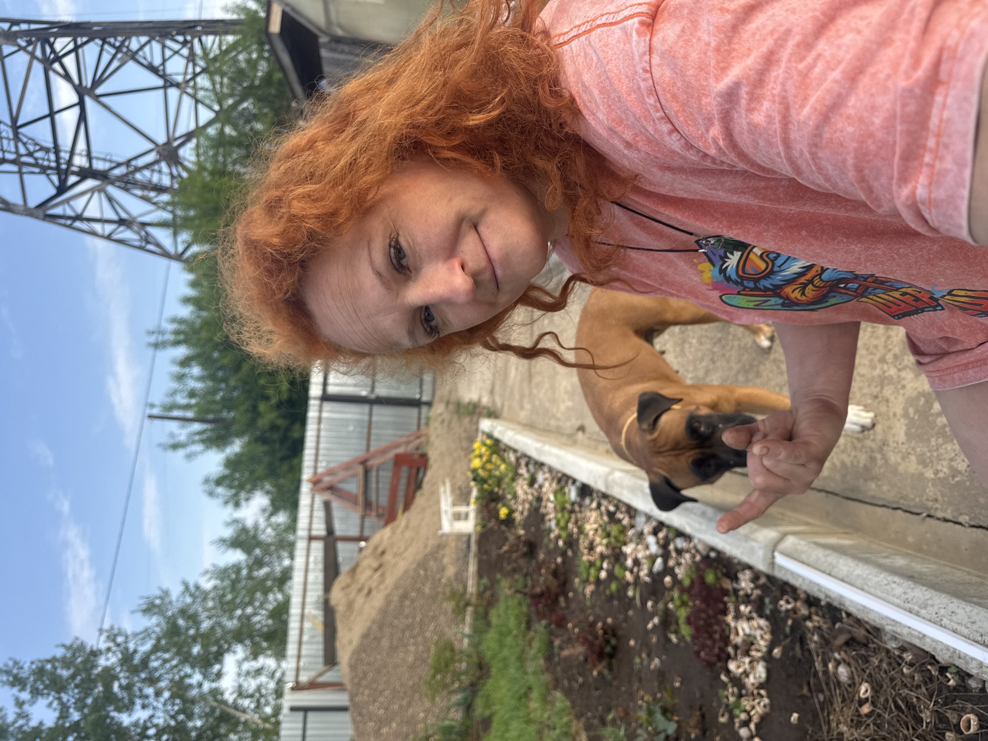 A selfie taken outdoors in Chaikovsky, Russia features a red-haired woman in a salmon-colored t-shirt with a graphic design. She is looking at the camera with a slight smile. A brown Boxer dog with black facial markings is positioned behind her, looking towards the woman's hand. The background includes a concrete sidewalk or patio area and a raised garden bed filled with rocks and plants. A gray metal fence, trees, and the base of a utility tower are visible in the background. The sky is partially cloudy.