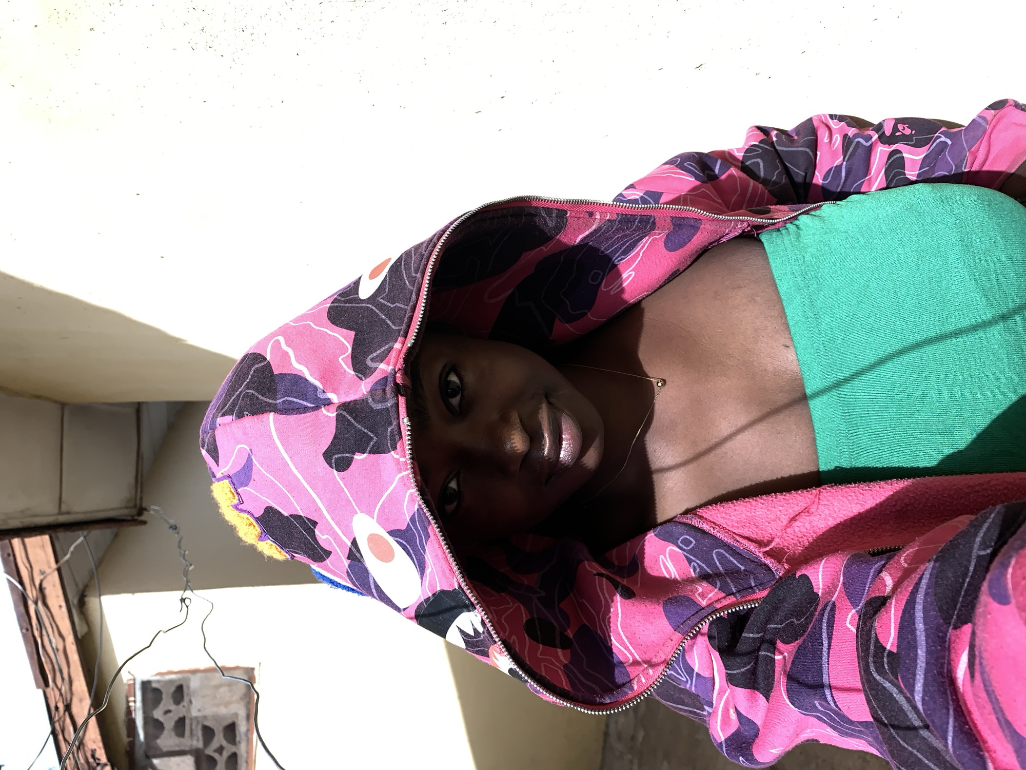 A woman is lying down, possibly on a surface. She is wearing a green top and a pink and purple camouflage hoodie. A gold necklace is visible around her neck. The background includes an exterior wall and some exposed wiring/metal structures, hinting at an urban or possibly semi-urban setting, which could be in Rafin Par, Nigeria. The lighting is bright, with strong shadows present.