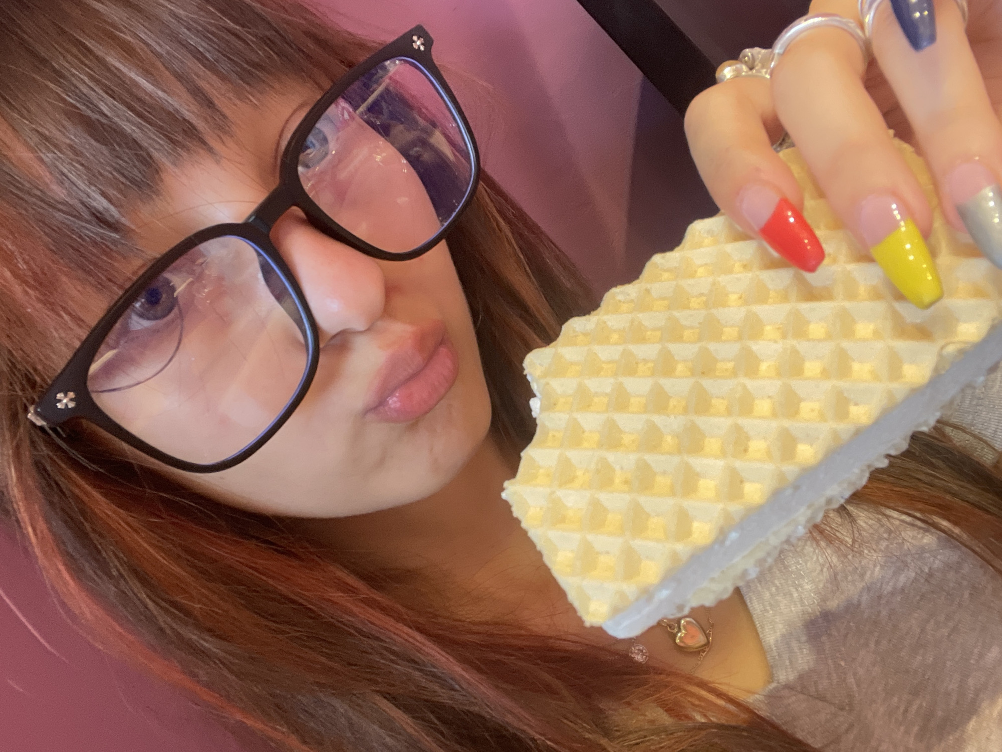 The image shows a close-up, indoor selfie of a person wearing black-rimmed glasses and holding a wafer snack. The person's hair is reddish-brown with some lighter streaks and bangs. They are wearing a grey top with a small heart-shaped pendant necklace. The wafer is rectangular with a grid pattern and appears to be filled with white cream. The person's right hand is visible, holding the wafer. Their fingernails are long and painted in a colorful design: red on the thumb, yellow on the index finger, and blue on the pinky. Several rings are visible on their fingers. The background is a solid, textured pinkish-purple color. The image appears to be taken in a domestic or casual setting. There is no information in the image to indicate it is located in Raanana, Israel.