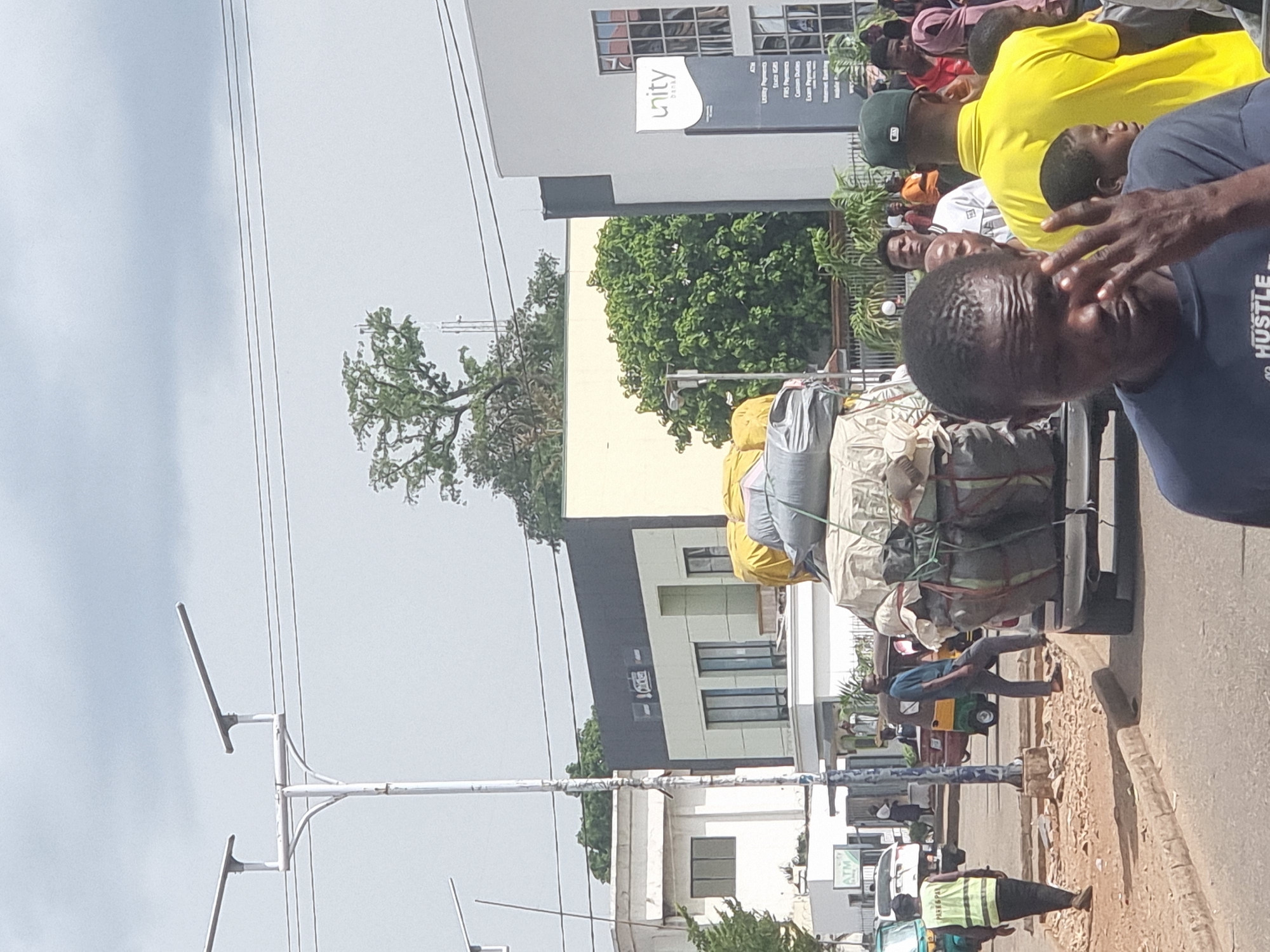 The image, likely taken in Jos, Nigeria, shows a busy street scene. A group of people is visible in the foreground, with one man wearing a blue shirt with "HUSTLE" printed on it. In the background, a commercial building is visible with a sign that reads "Unity". The building has multiple windows and appears to be modern in design.
A vehicle, heavily loaded with bagged items, is also in the frame. Some of the bags appear to be yellow and gray. The vehicle is surrounded by people, suggesting a congested area. The sky is overcast, and electrical wires stretch across the top of the frame. A white lamppost is also visible. A tricycle taxi is seen in the road. The general impression is of a bustling commercial area with people engaged in various activities.