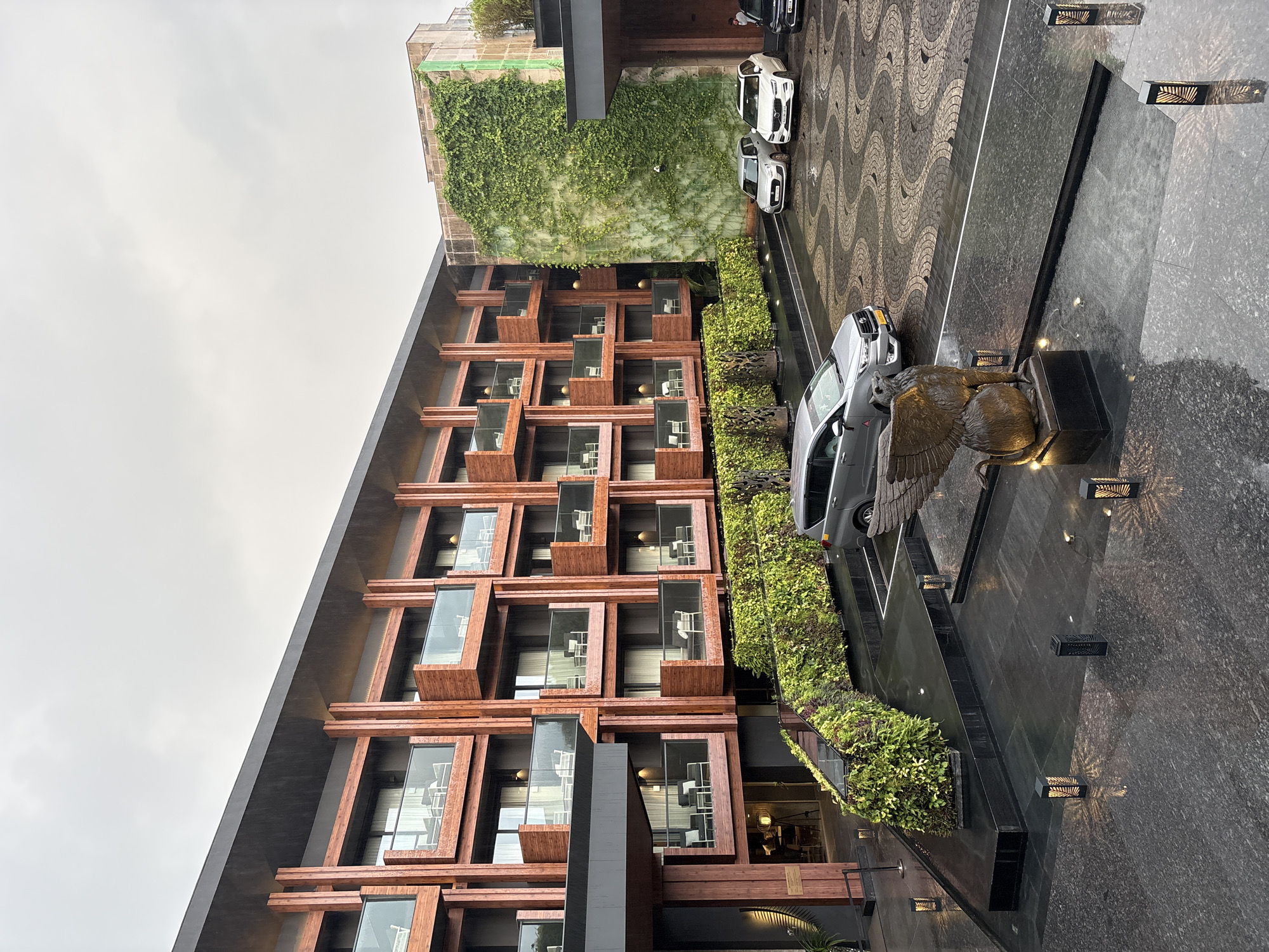 An exterior shot captures the facade of a modern hotel, likely in Vagator, India, under a cloudy, overcast sky with visible raindrops. The building features a contemporary design with balconies, each accented by wooden beams and glass railings. Green, leafy plants adorn the exterior walls, creating a striking vertical garden. In the foreground, a paved area with a patterned design shows several cars parked, including a silver SUV, a white SUV, and a black sedan. A sculpture of a winged creature stands prominently in the foreground. The ground appears wet, suggesting recent rain. The overall impression is one of an upscale, nature-integrated hotel establishment.