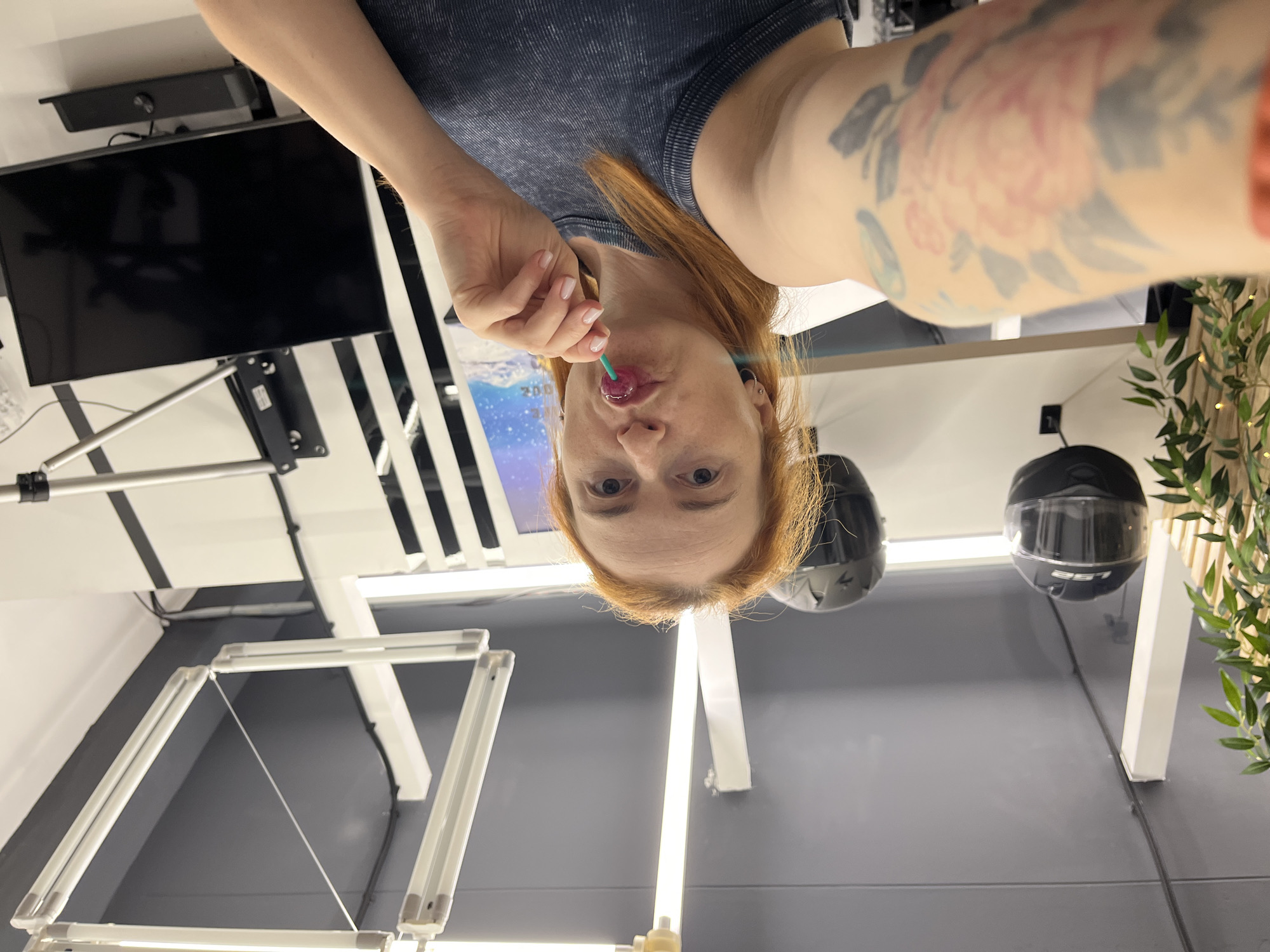 A woman with auburn hair, wearing a dark grey ribbed tank top, is taking a selfie. She has a lollipop in her mouth and is holding it with her right hand. She has a tattoo of a pink flower on her left arm. In the background, there are two black helmets hanging on the wall, a plant with green leaves, fluorescent lights on the ceiling, and a black television screen mounted on the wall. The scene appears to be indoors, possibly a workshop or a studio.