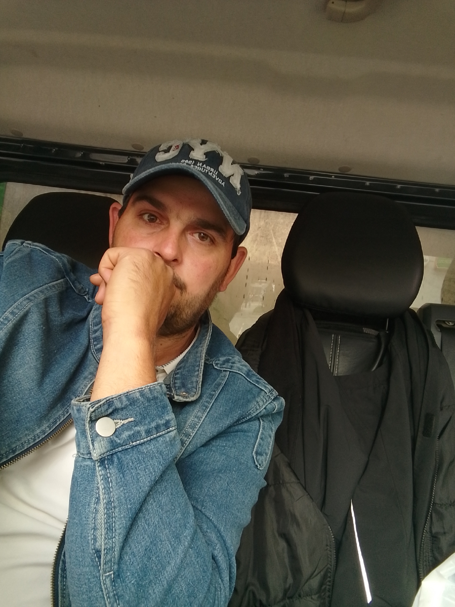 The image shows a man in the driver's seat of a vehicle, likely a car or truck. He is wearing a blue denim jacket and a baseball cap. His right hand is resting on his chin, and he is looking directly at the camera. The interior of the vehicle is visible, with a black headrest and a black jacket hanging on the side. The scene is indoors and appears to be during the daytime. There is no specific indication of the city of Pinneberg, Germany, within the image itself.