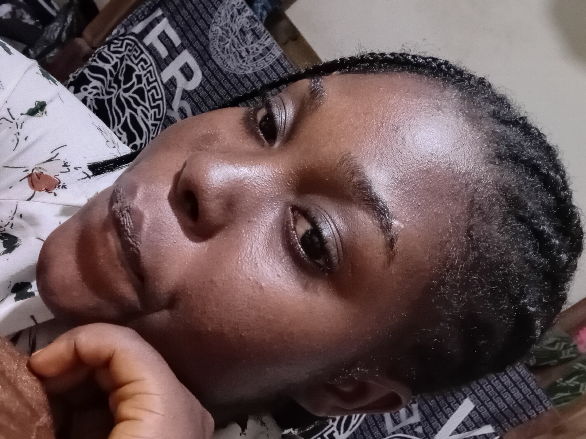 The image shows a close-up, head-and-shoulders view of a young Black woman lying down, with her head tilted. She has her eyes open, looking towards the camera. Her hair is styled in braids, and she is wearing a white top with a floral print. The background includes a patterned fabric, possibly a bedsheet or pillowcase, with a repeating geometric design and the letter "F" visible on some sections. The lighting suggests an indoor setting. The image is not cropped to indicate a specific location in Ejule, Nigeria.