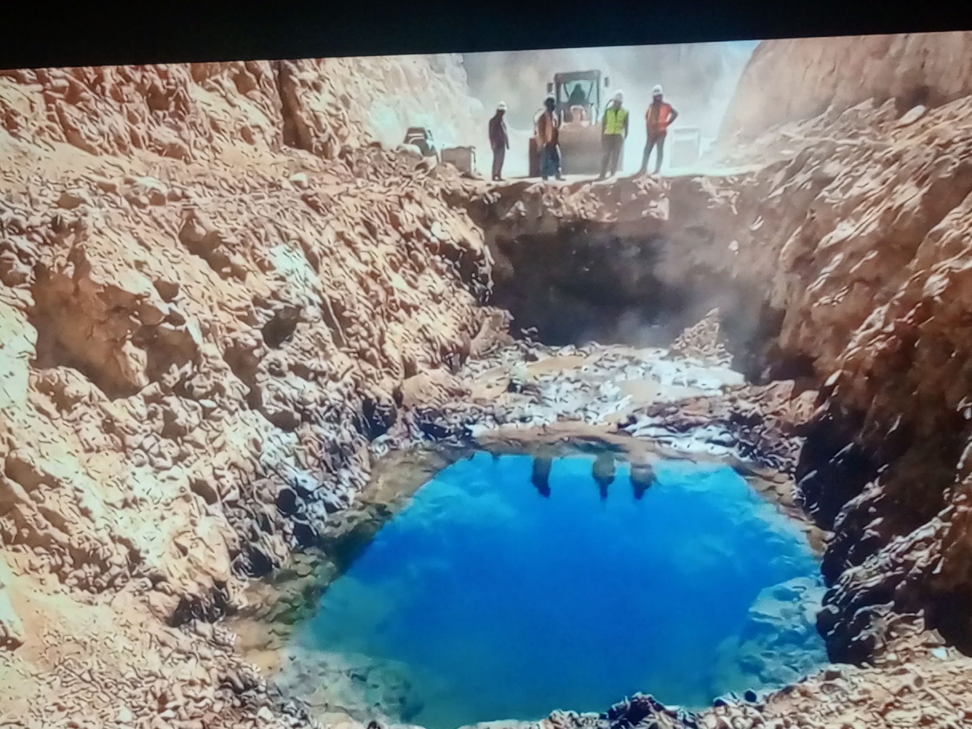 A high-angle, eye-level shot captures a surreal, bright blue pool of water in the middle of a dusty, rocky quarry. The pool is roughly circular and appears to be an unexpected feature within the otherwise barren landscape. The surrounding terrain is characterized by steep, eroded rock formations in shades of tan and brown, with loose debris scattered at the edges of the pool.
At the rim of the quarry, near the top of the image, a group of individuals are visible. They are standing on the edge of the disturbed earth. One person, wearing a white hard hat and an orange vest, stands facing away from the viewer. To their left, another person in a white hard hat and a yellow vest is also facing away. Further left, a person in a dark shirt and white hard hat stands, looking towards the pool. In the background, a yellow construction vehicle, possibly a bulldozer or front loader, is parked. Another smaller, lighter-colored construction vehicle is also visible.
The water in the pool is a vibrant, almost unnatural shade of blue, reflecting the sky or possibly the composition of the water itself. The edges of the pool are rocky and uneven, suggesting it has formed within the quarry. There is a haze or dust rising from the quarry floor, obscuring some of the details further into the excavation. The overall impression is one of a man-made excavation site that has encountered an unusual natural phenomenon or a man-made reservoir in an unexpected location.