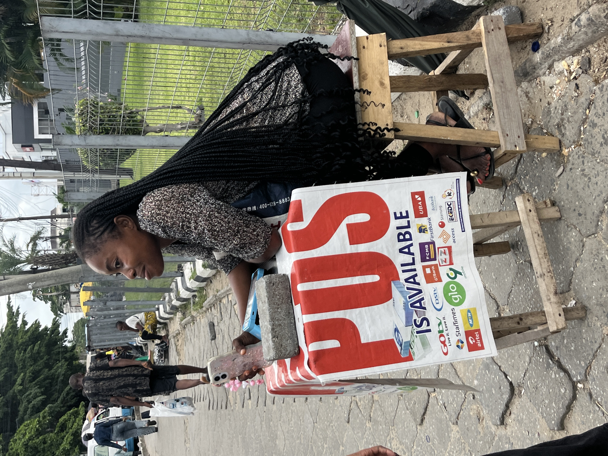 A woman with long braided hair sits on a wooden stool beside a sidewalk in Itirin, Nigeria. She is leaning over a makeshift table covered with a sign that reads "POS IS AVAILABLE". The sign also displays logos of various Nigerian telecommunications and banking companies. She is holding a cell phone in one hand and a brick or concrete block rests on the table. The sidewalk is cracked and worn. In the background, other people walk along the sidewalk, and cars drive on the street. A wire fence separates the sidewalk from a green lawn and buildings.