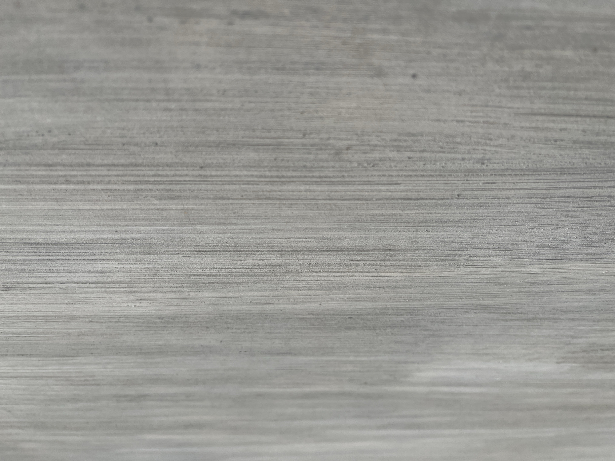 The image is a close-up, vertical shot of a textured, gray surface. The texture is characterized by numerous fine, parallel lines running from top to bottom, suggesting a material like brushed concrete, wood grain, or a painted surface with visible brush strokes. There are subtle variations in shade and tone, with some areas appearing lighter and smoother, while others have darker, more pronounced lines and occasional small specks. The lighting creates a slight shadow effect along the vertical lines, emphasizing the texture. The image does not contain any people, animals, or discernible objects other than the surface itself. There is no visible context indicating a specific location within Teluk Air, Indonesia.