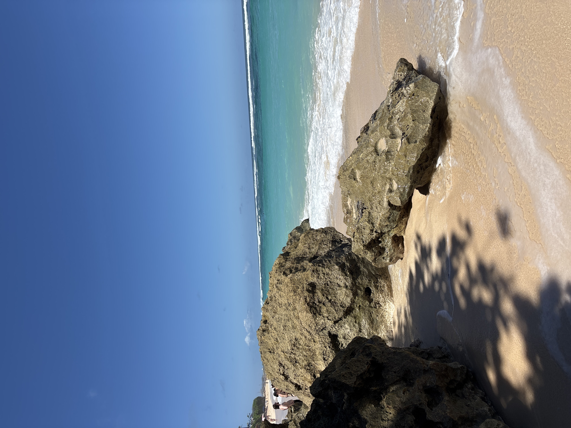 The image depicts a coastal scene in Kuta Selatan, Indonesia. The sky is a clear, bright blue. The turquoise ocean meets the sandy beach, with white waves gently rolling in. Large, textured rocks are present in the foreground, partially obstructing the view of the beach. A shadow from foliage overlays the sand near the rocks. In the distance at the base of the cliff, a few people and buildings are visible.