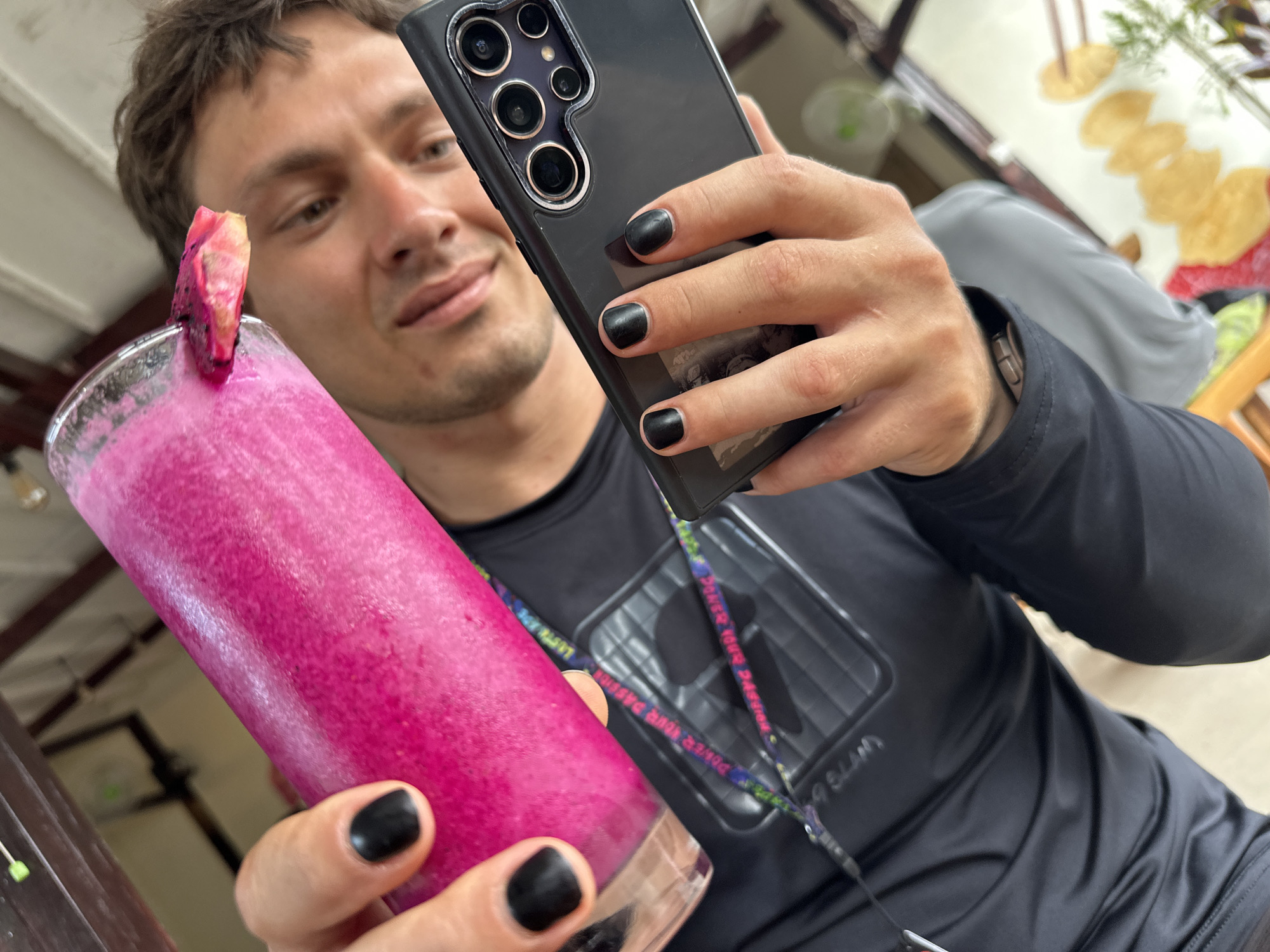 A person with short brown hair, light skin, and painted black fingernails is taking a selfie with a black smartphone. The phone has multiple camera lenses on the back. In their left hand, they are holding a tall glass of a bright pink beverage, garnished with a slice of dragon fruit. The person is wearing a black athletic shirt with a lanyard around their neck. In the background, a blurred environment suggests an indoor or covered outdoor setting, possibly a cafe or restaurant. The location is indicated as Unknown, Indonesia. Other details include the presence of a table and someone else seated at the table to the right of the person taking the picture.