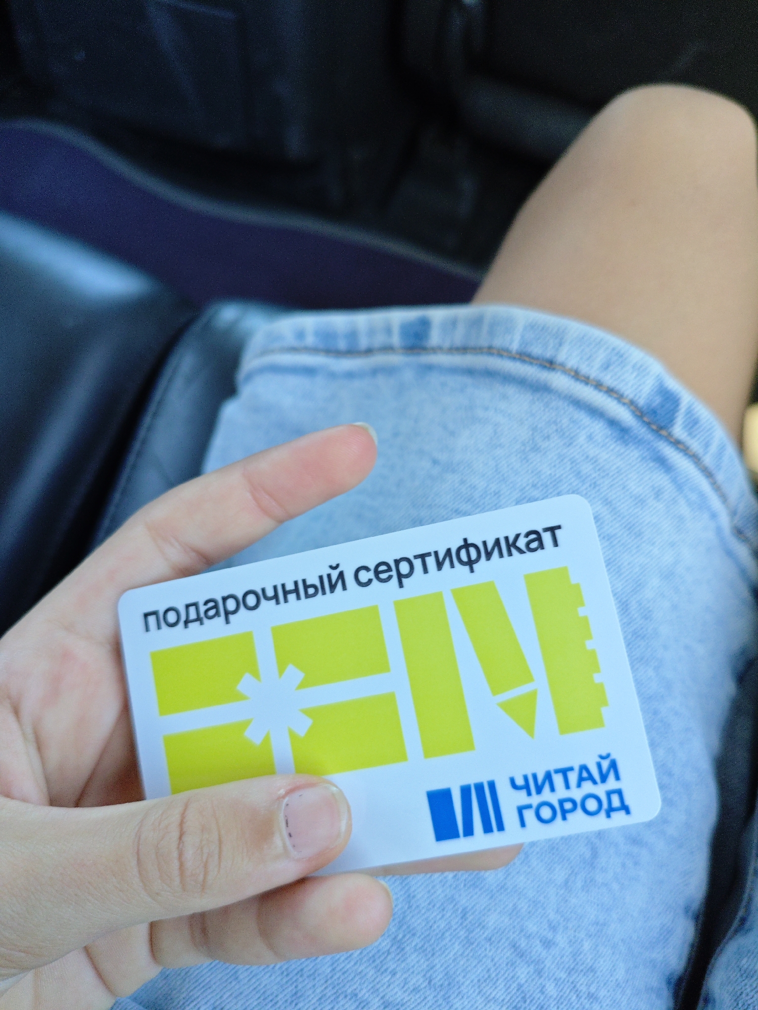 A hand is holding a gift certificate with the text "подарочный сертификат" (gift certificate) printed in Russian. The certificate features a yellow and white design, including what appears to be a stylized gift box with a bow and a pencil. Below this, the logo "ЧИТАЙ ГОРОД" (Read City) is visible in blue letters, accompanied by a blue graphic of vertical bars. The certificate is held over a person's lap, which is covered by light blue denim shorts. The background is blurred, showing dark upholstery, possibly of a car seat. The scene is not specifically indicative of the city of Трудовское сельское поселение, Ukraine, as there are no identifiable landmarks or contextual clues present in the image.
