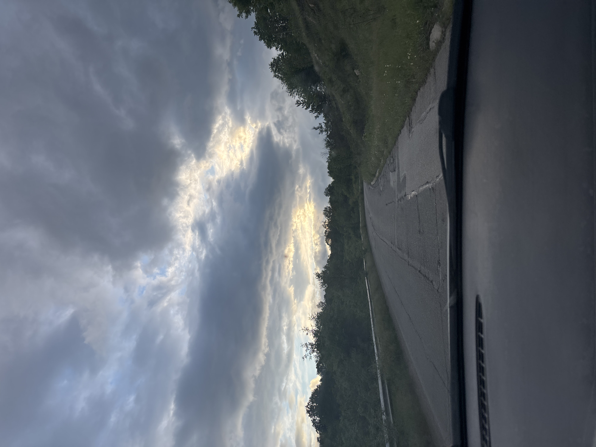The image depicts a view from inside a vehicle traveling on a narrow road in Golema Rakovitsa, Bulgaria. The road curves slightly to the right, lined with green vegetation and trees on one side. The sky is filled with dramatic clouds, with hints of sunlight breaking through. Part of the vehicle's interior is visible on the right side of the frame.