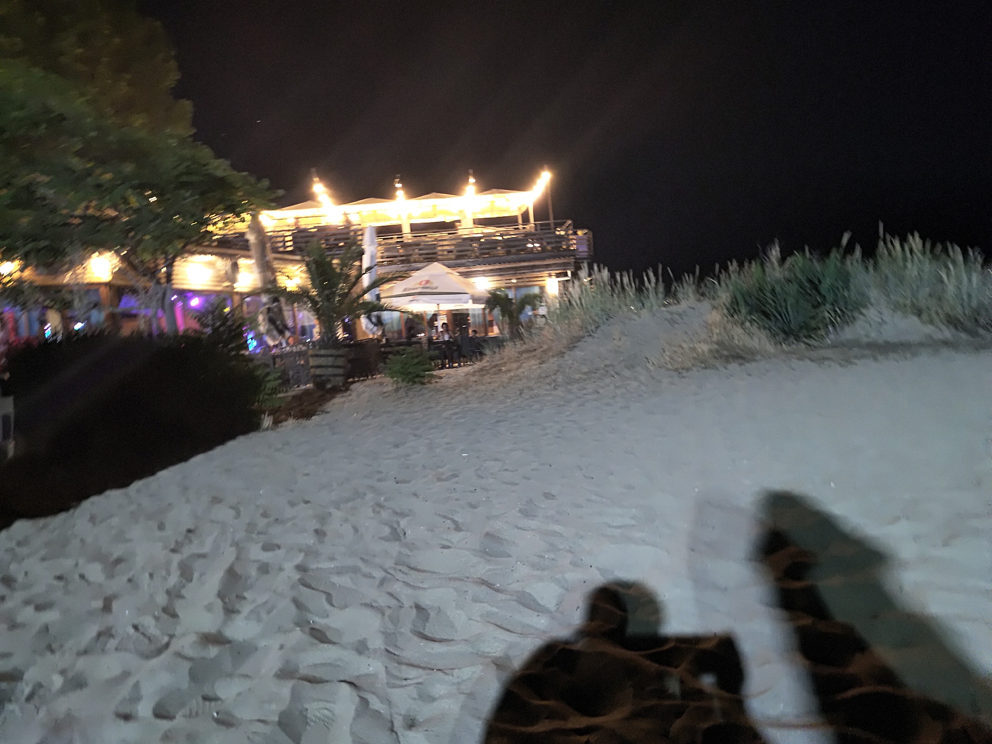 A night-time scene captures a sandy beach with a tiered building illuminated by lights in the background. The building appears to be a restaurant or bar, with multiple levels and outdoor seating. A large tree with foliage is visible on the left side of the frame, and tall grasses grow on a sandy dune to the right. The foreground is dominated by footprints in the sand, and shadows of people can be seen on the beach. The sky is dark, indicating it is nighttime. The scene suggests a lively atmosphere at a beachfront establishment.