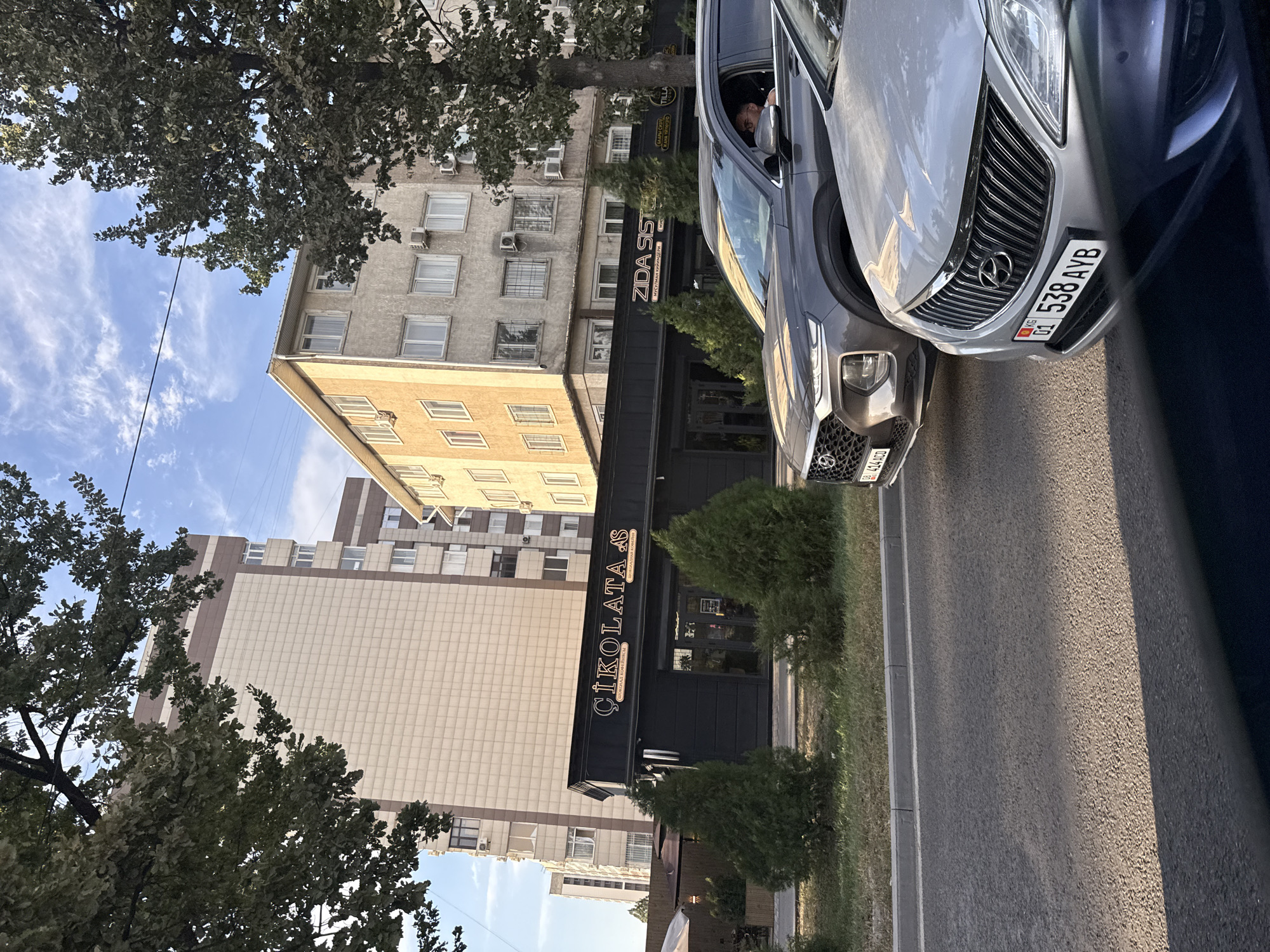 This is a street-level photograph taken in Bishkek City, Kyrgyzstan, focusing on a storefront and the street. In the foreground, two cars are visible, the closest being a silver sedan with a prominent chrome grille and a license plate reading "01 538 AYB KG". Behind it, a dark gray SUV is partially visible, with its front grille and a portion of its driver's side visible. The driver, a man wearing glasses, is partially seen through the window.
The middle ground features a black storefront with illuminated white lettering that reads "ÇIKOLATA AS" above smaller text in a different language. Adjacent to this storefront is another sign reading "ZIDA SISTERS" in a similar font. The buildings behind the storefront are multi-story residential or commercial structures, one appearing lighter in color with a grid pattern and the other a muted yellow with numerous windows. Several coniferous trees are planted in front of the storefront, adding greenery to the urban landscape. The sky above is blue with scattered white clouds, and sunlight casts shadows on the asphalt street. Power lines are visible crisscrossing the sky.