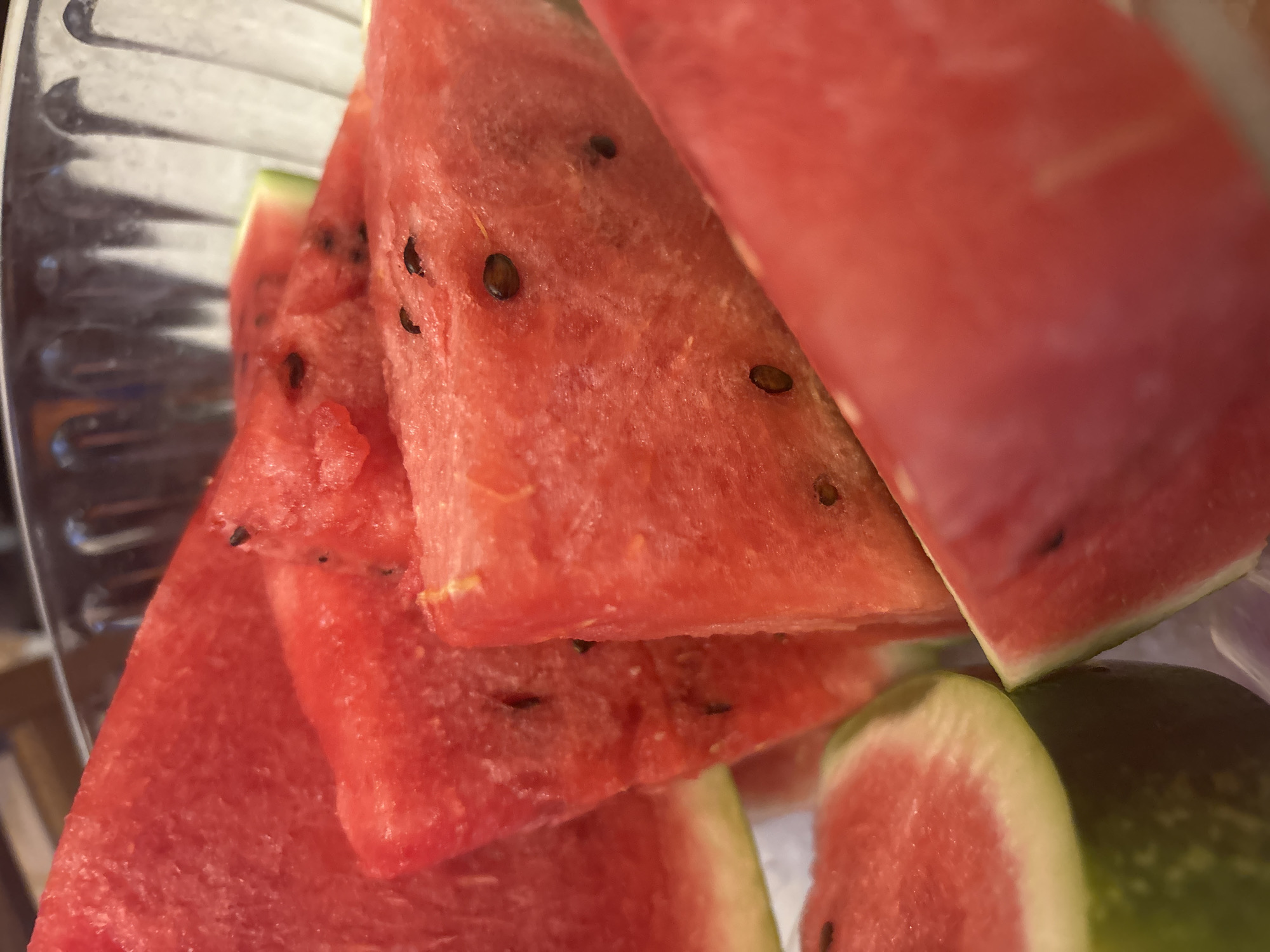 A close-up, overhead shot shows several slices of watermelon in a clear glass bowl. The watermelon is bright red with black seeds visible on the flesh of some slices. The rind is green and white. The slices are arranged in a slightly overlapping manner, filling most of the bowl. The lighting is bright, highlighting the juicy texture of the fruit. The background is blurred, but appears to be a wooden surface. There are no people or animals in the image. The location is not determinable from the image content, and therefore cannot be specified as Trudovets, Bulgaria.
