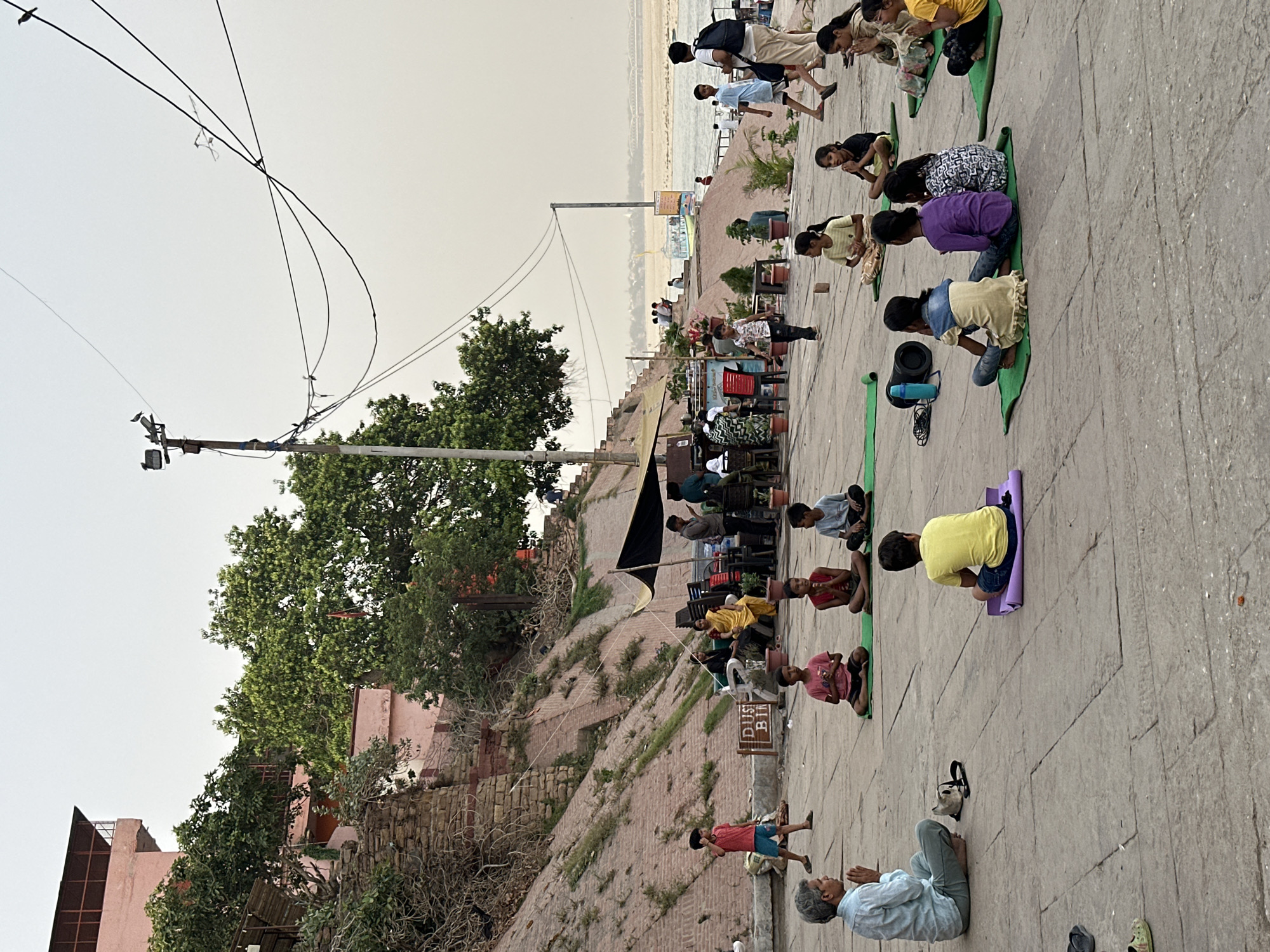 The image shows an outdoor yoga class taking place in Varanasi, India. Several children are participating, seated on yoga mats, while an adult sits nearby with their hands in prayer position. A "Dust Bin" sign is visible. In the background, there is a raised area with trees, a building with a tented area, and other people in the distance near the river. Overhead are electrical lines.
