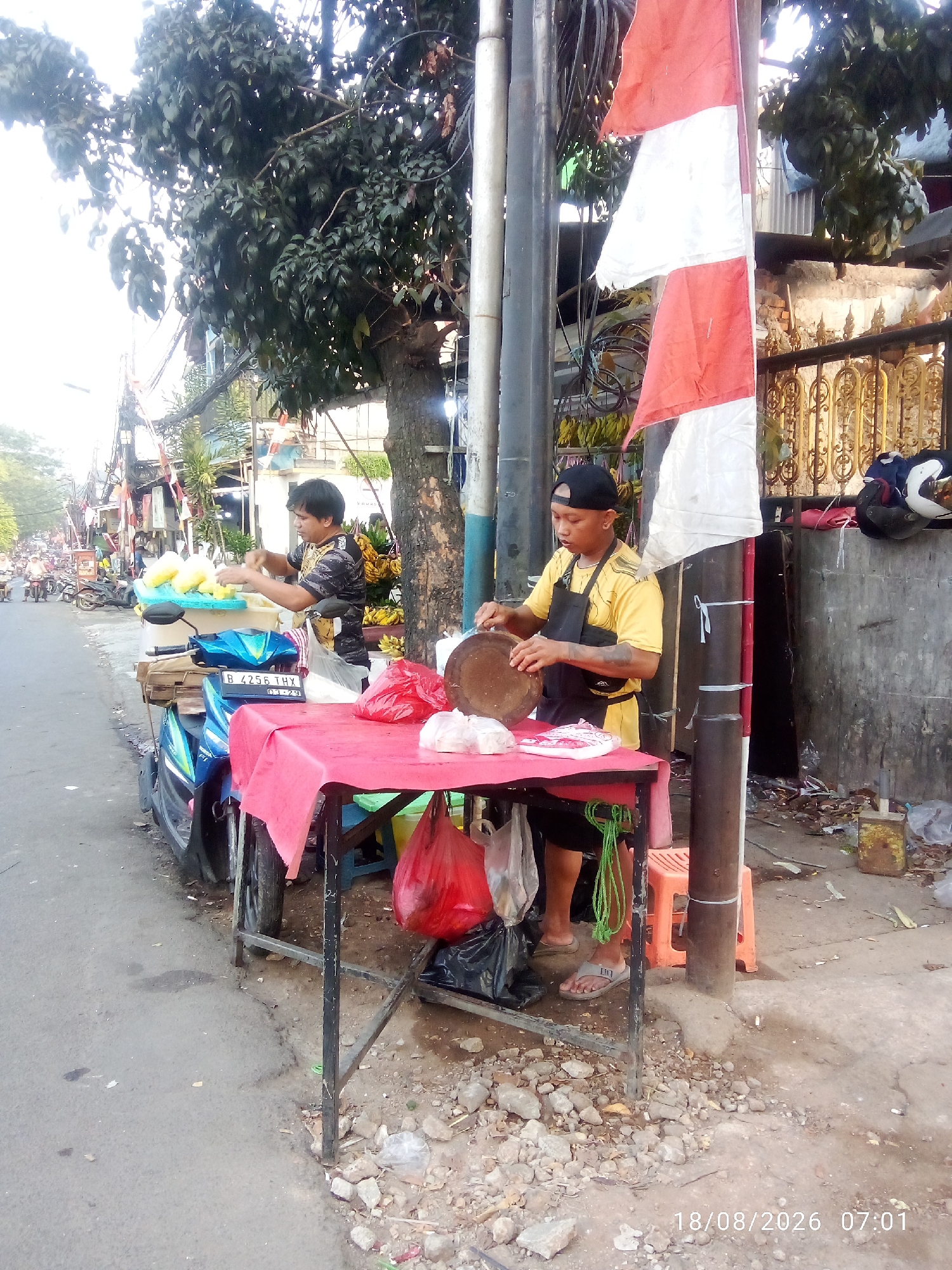A street vendor is preparing food on the side of a road in East Jakarta, Indonesia. A man, wearing a yellow t-shirt and a black apron, stands behind a table covered with a red tablecloth. He is holding a circular cutting board and appears to be slicing something. Next to him, another person, possibly a customer or helper, is arranging yellow fruits in a blue container on a blue motorcycle. The street is lined with trees, utility poles with many wires, and parked motorcycles. A red and white Indonesian flag is displayed on a pole. In the background, there are more vendors and traffic, suggesting a busy market area. The ground is paved with asphalt, and there are rocks and debris on the side of the road. The timestamp on the image indicates August 18, 2026, at 07:01.