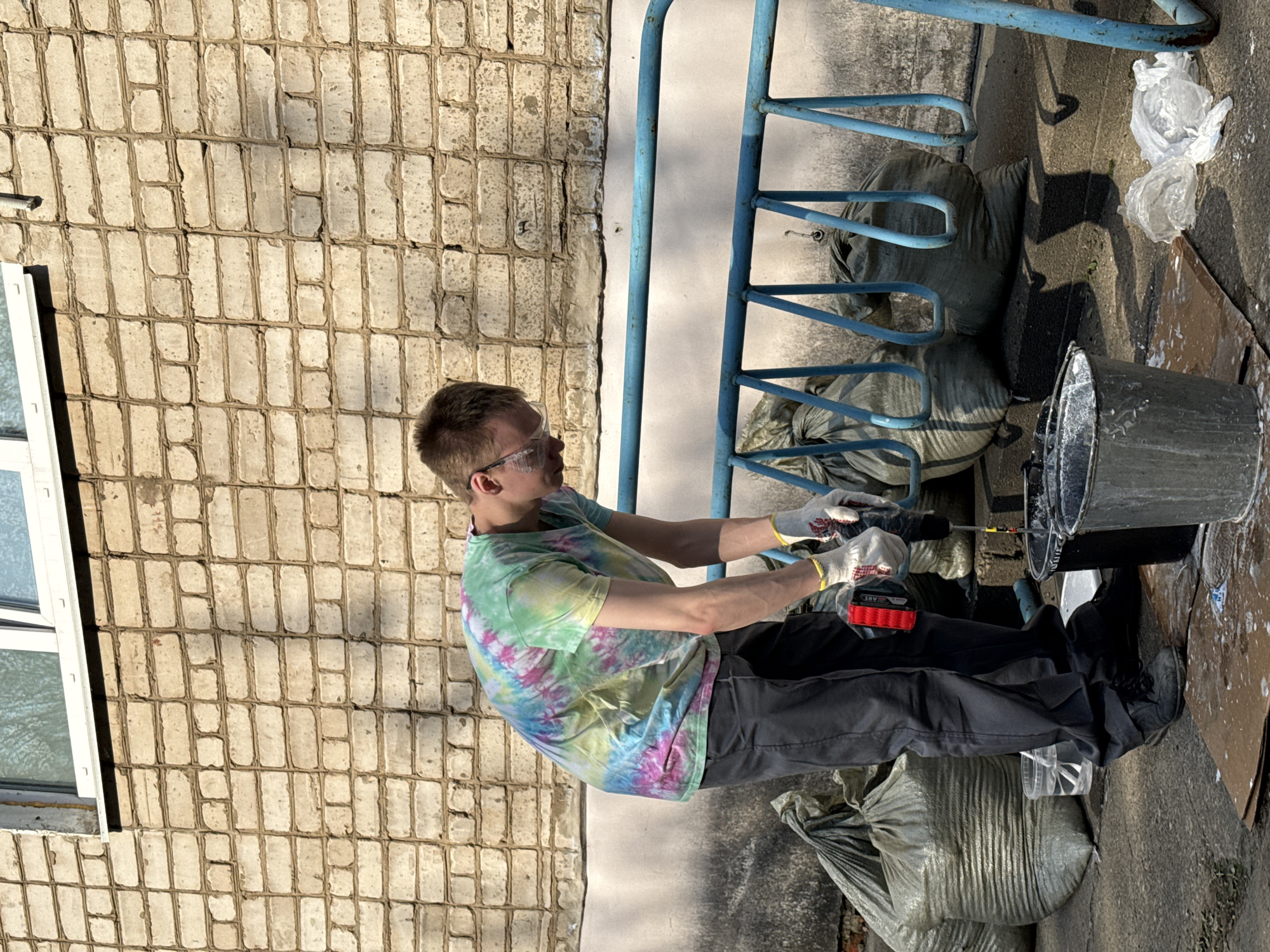 The image shows a man wearing safety glasses, gloves, and a tie-dye shirt using a power drill to mix something in a metal bucket. He is standing outside next to a brick building and a blue metal bike rack. There are several large sandbags and a plastic bag nearby. The location is Chaikovsky, Russia.