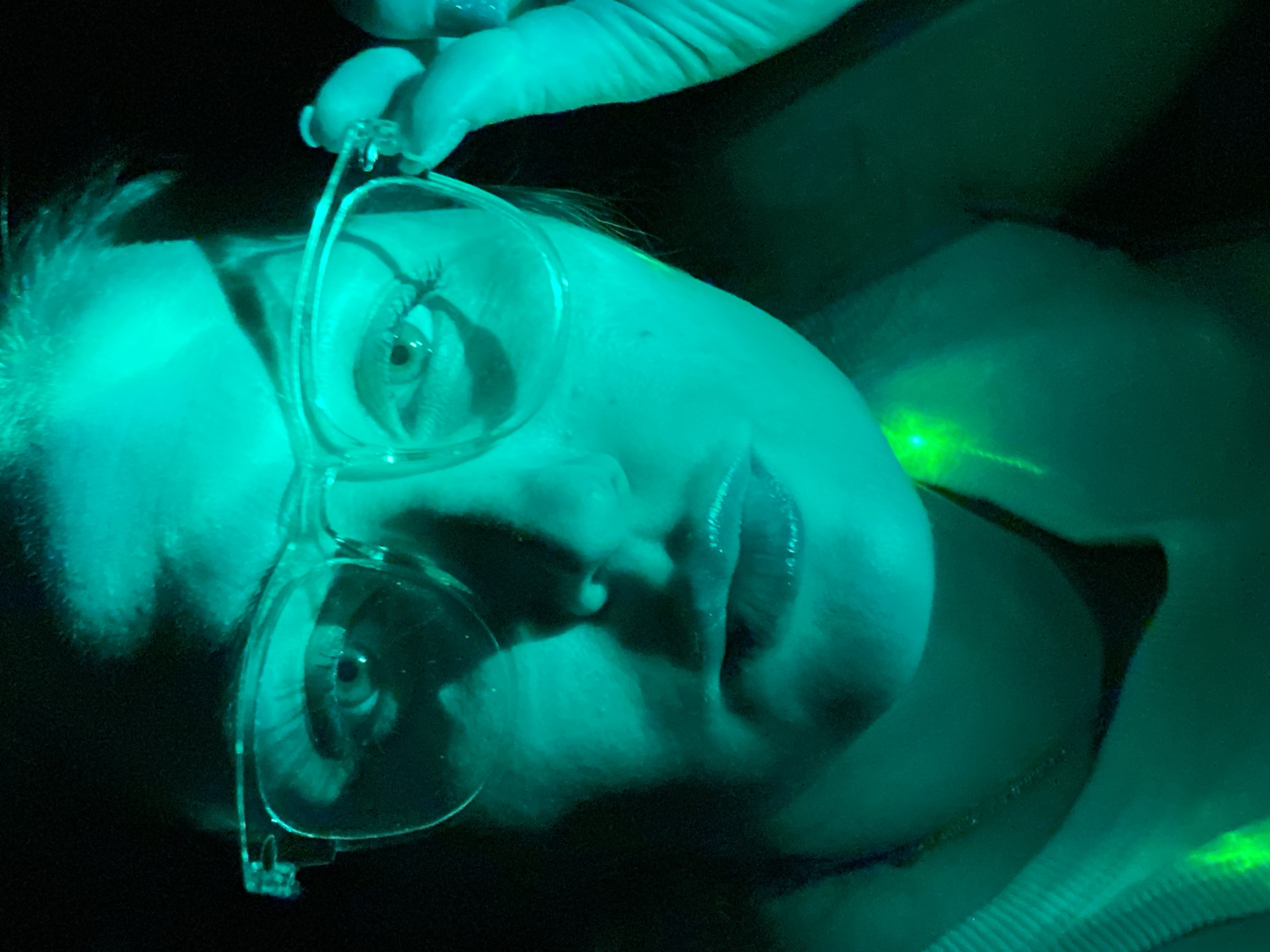 A face is visible from a low angle in dark lighting. The face appears to be that of a fair-skinned person wearing clear-framed eyeglasses. A hand is also visible, holding the eyeglasses. There is green lighting in the scene, casting a green hue over the face and glasses. No landmarks or elements within the image suggest the location of Марковский, Russia.
